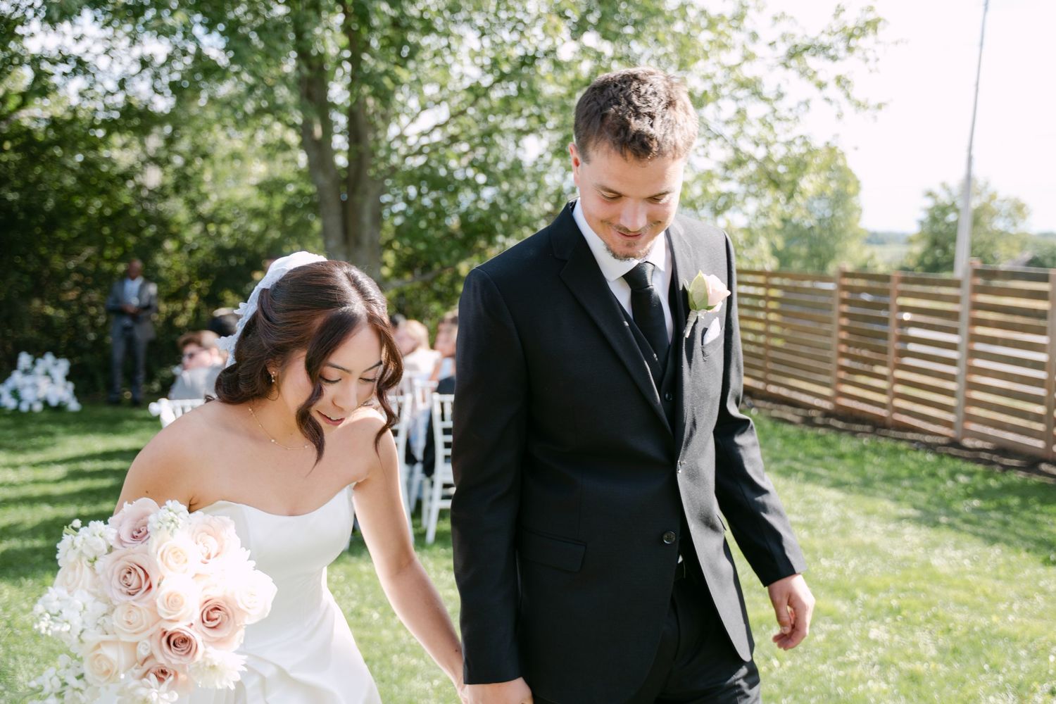 Smiling bride in a white gown holding a pink rose bouquet walks hand in hand with a groom in a black suit at an outdoor garden wedding.