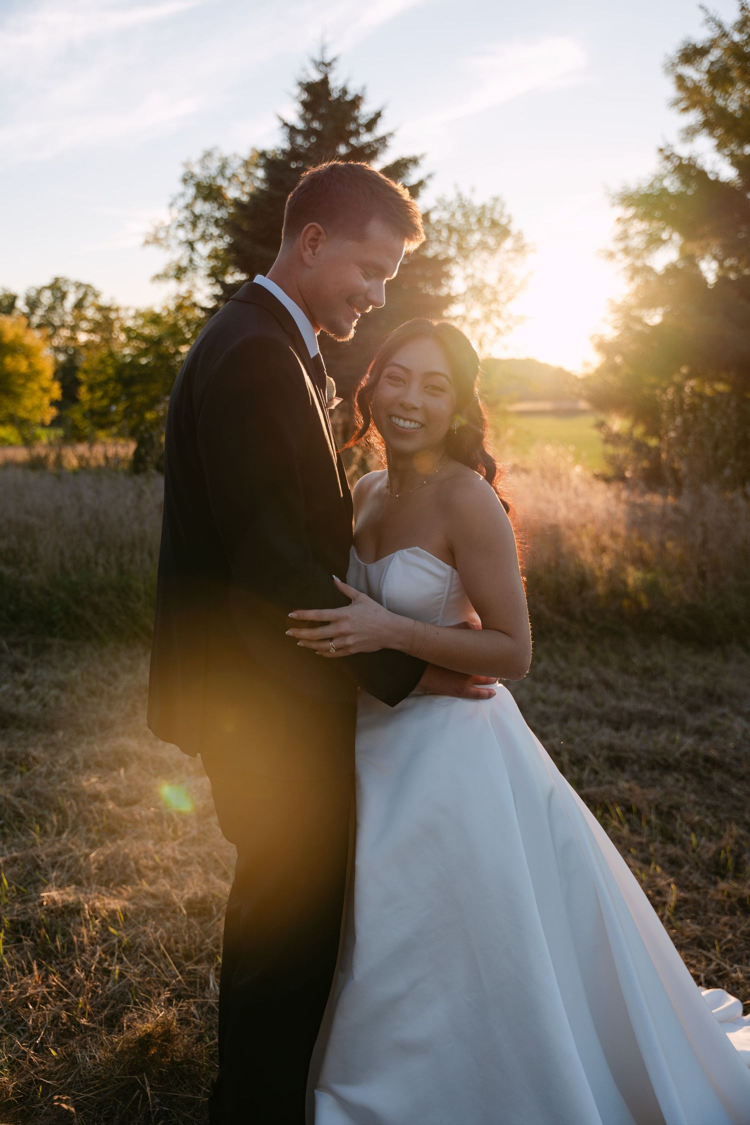 Smiling bride in a white strapless gown embraces her groom in a dark suit in a golden field at sunset on their wedding day.