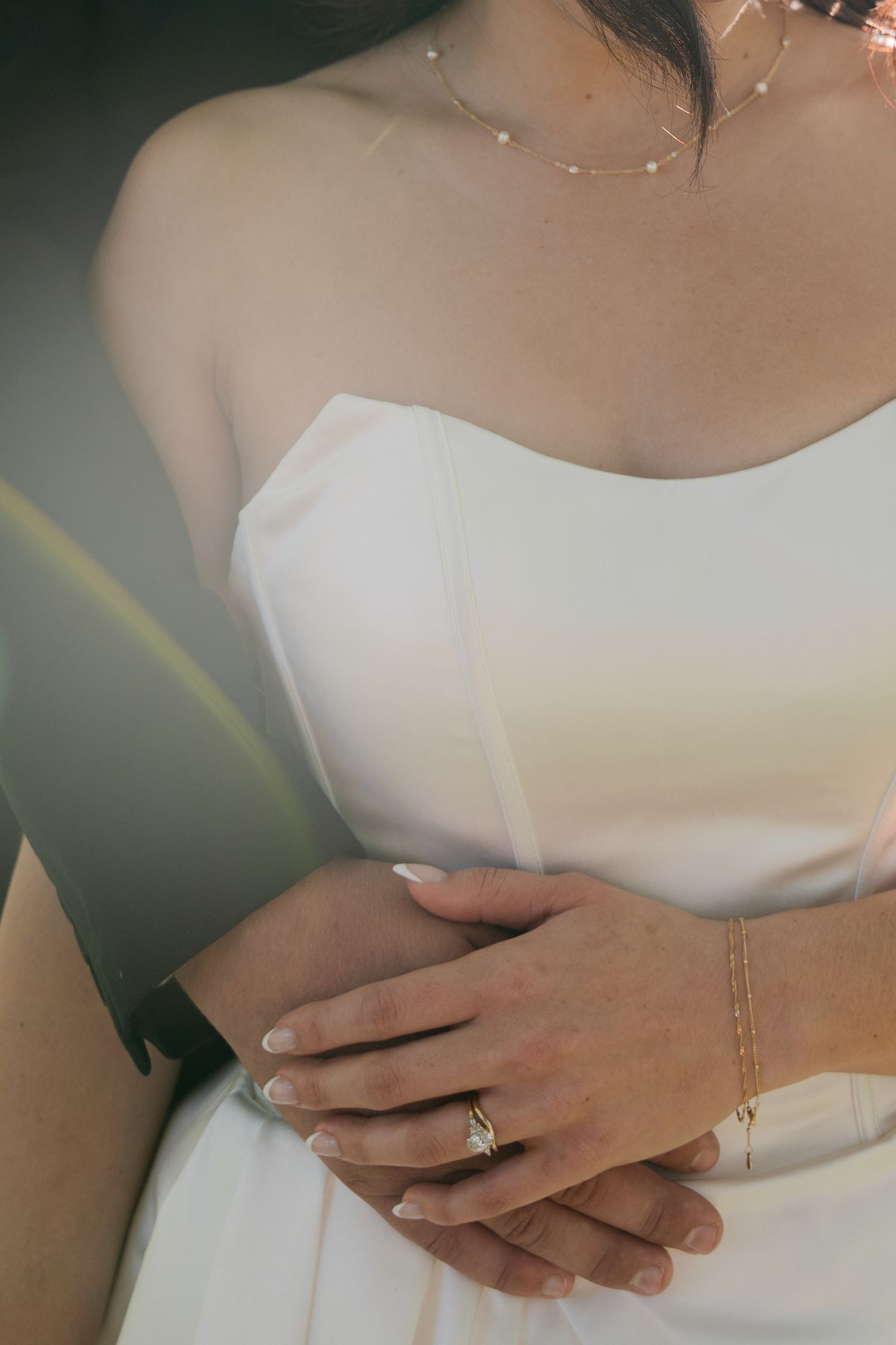 Bride in a strapless white satin gown wearing a diamond ring and gold bracelet as a groom in a dark suit wraps his arms around her.