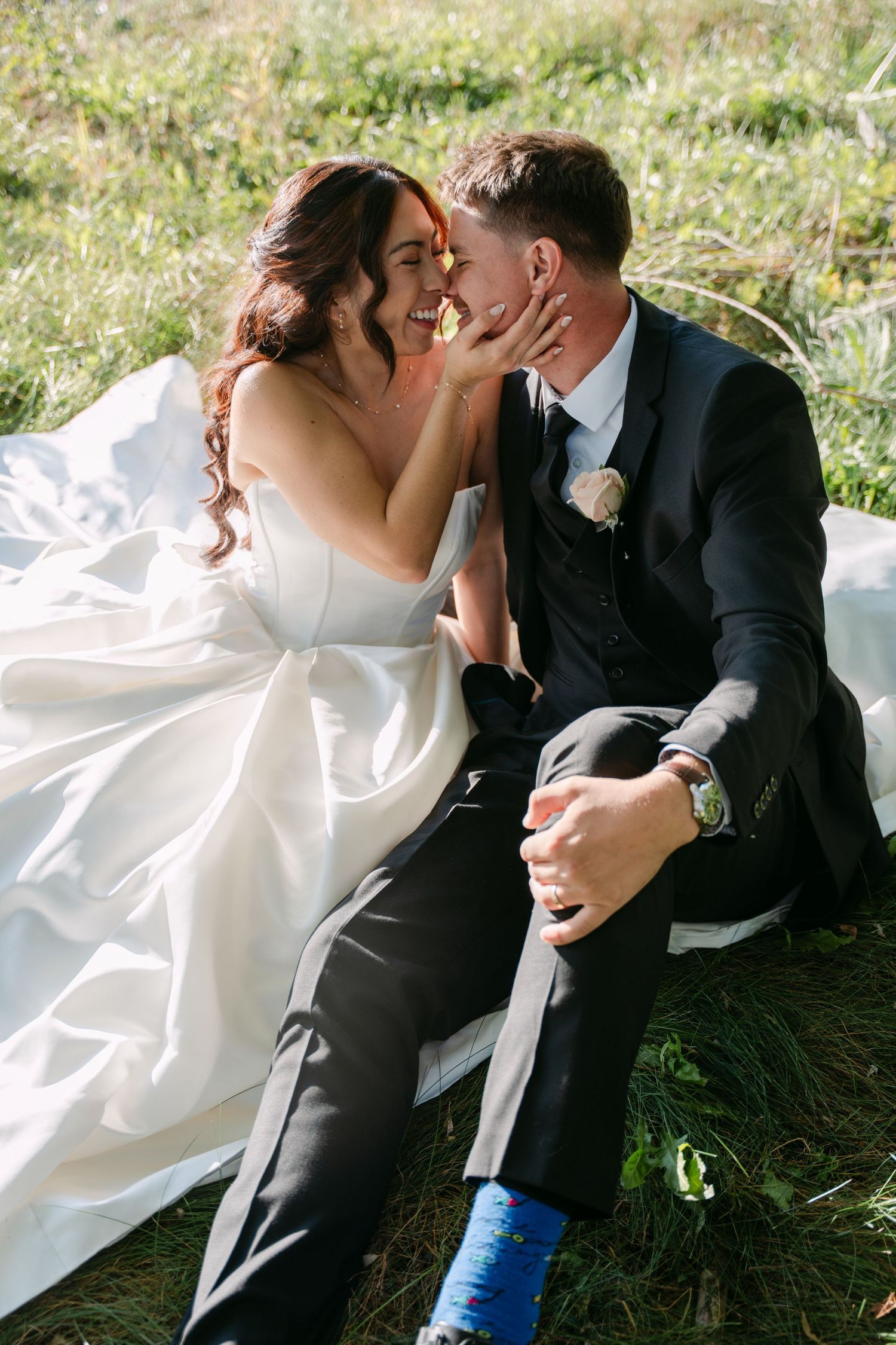 Smiling bride in a white satin wedding gown touches her groom's face as the couple sits close together on the grass.