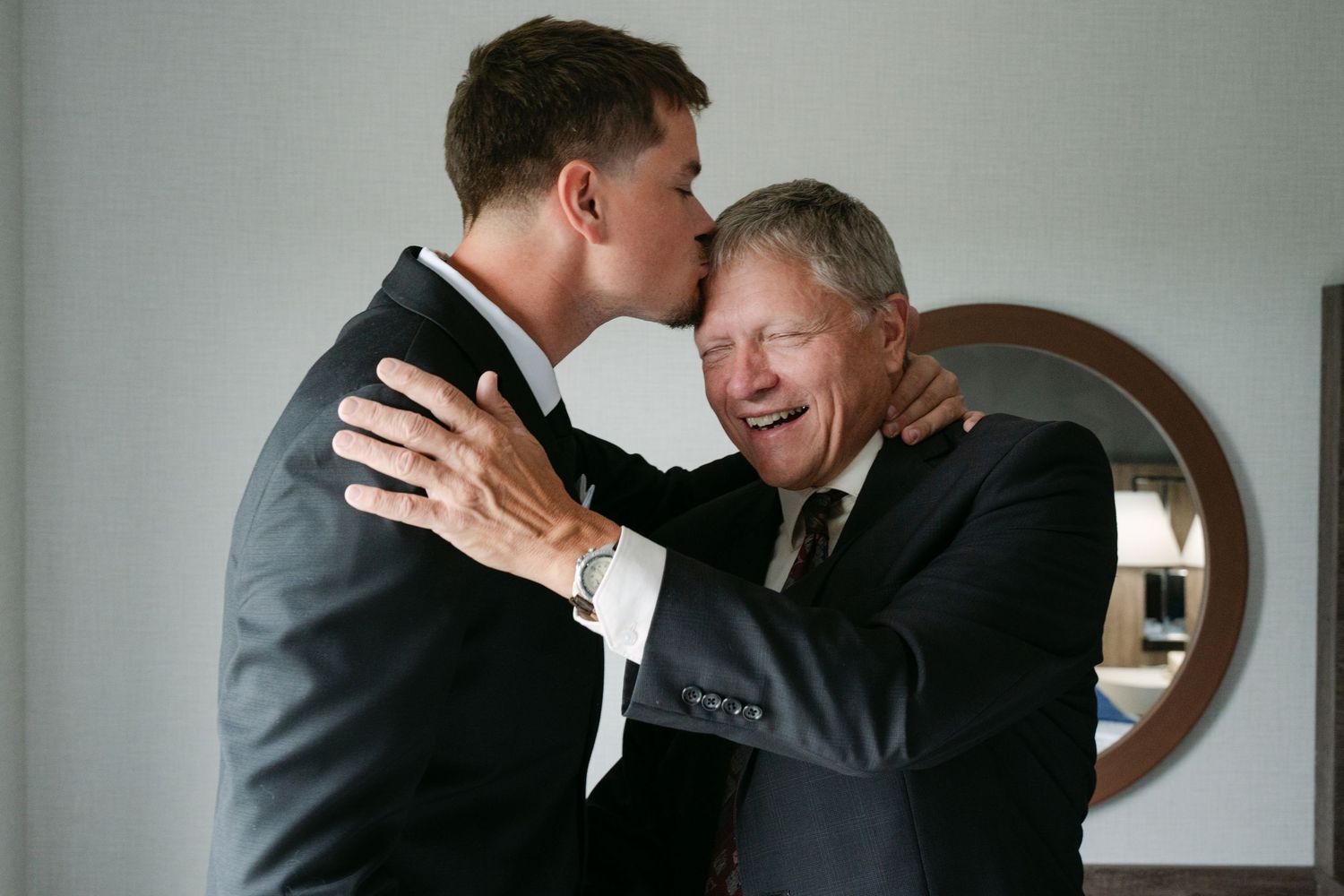 A young man in a dark suit tenderly kisses the forehead of a smiling older man in formal attire during an emotional moment indoors.