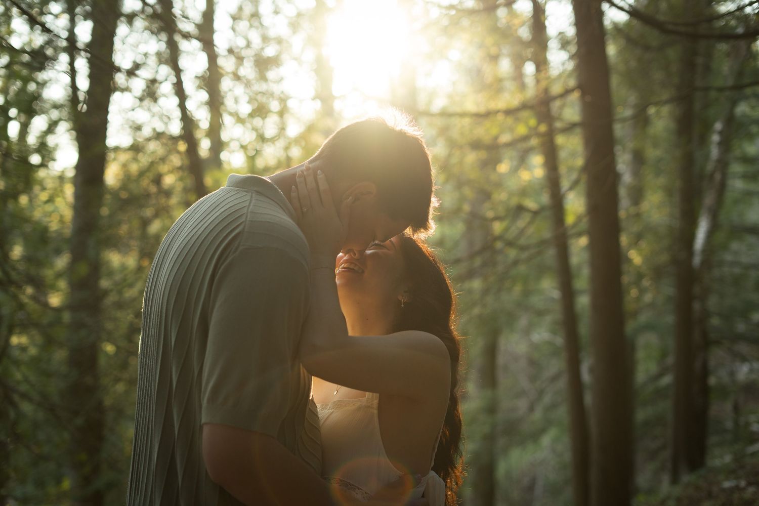 Couple sharing a romantic kiss in a sunlit forest during golden hour.