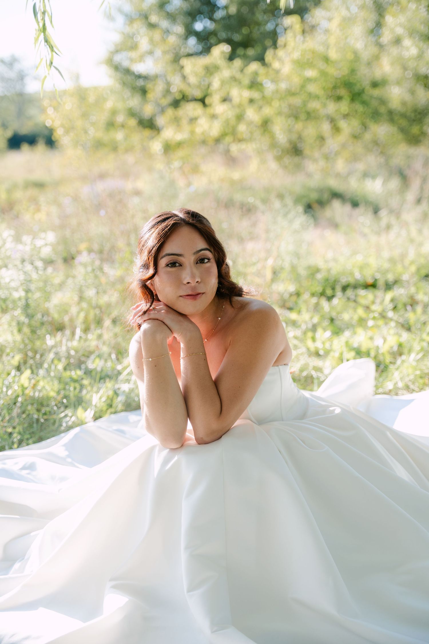 A bride in a strapless white satin ball gown rests her chin on her hands while seated in a sunlit green meadow.