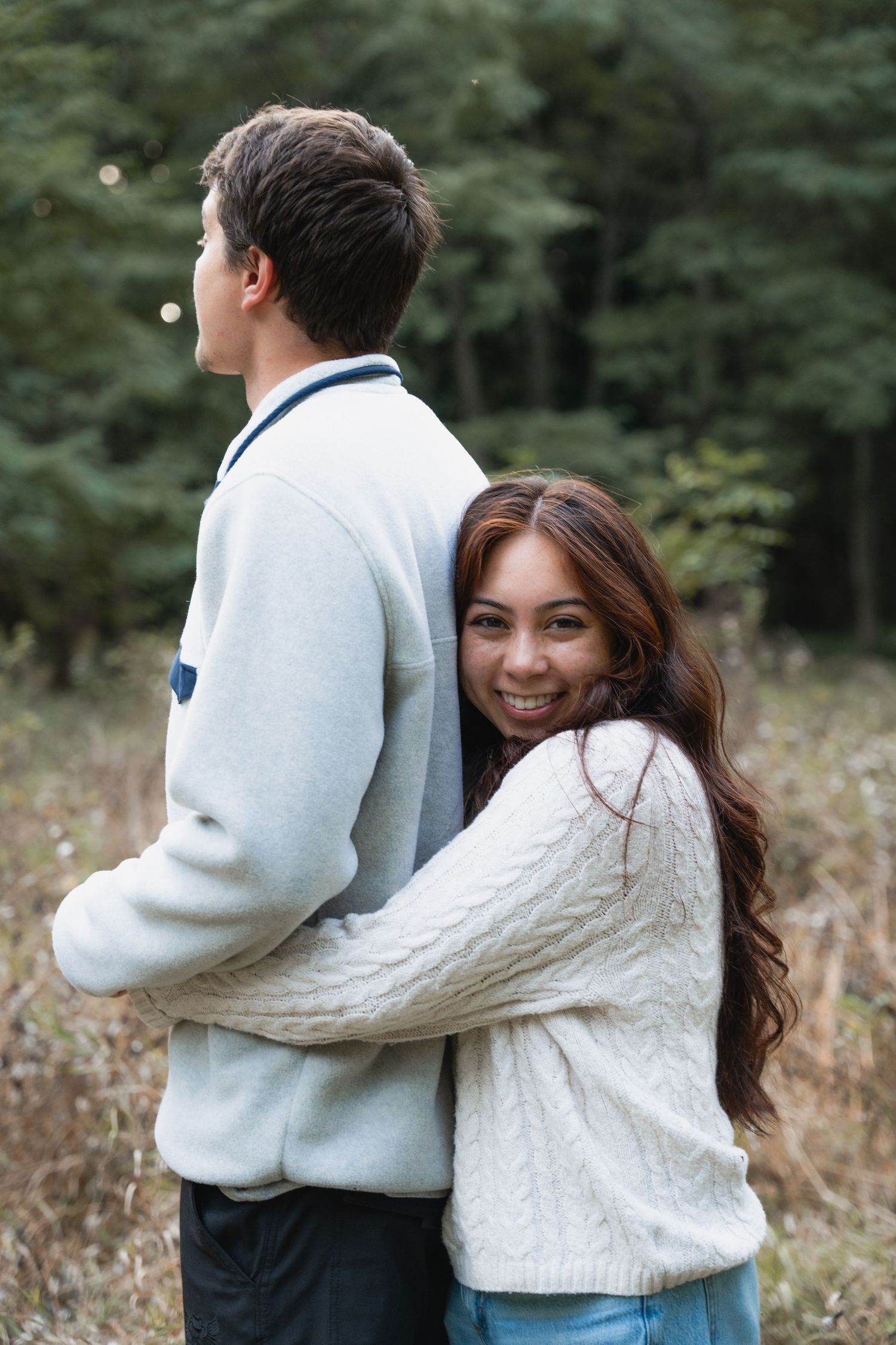 A couple in cozy sweaters embraces outdoors in a wooded setting.