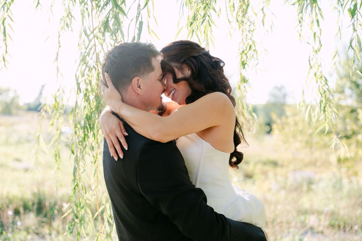 Bride in a white dress and groom in a dark suit embrace tenderly under weeping willow branches in a sunlit field.