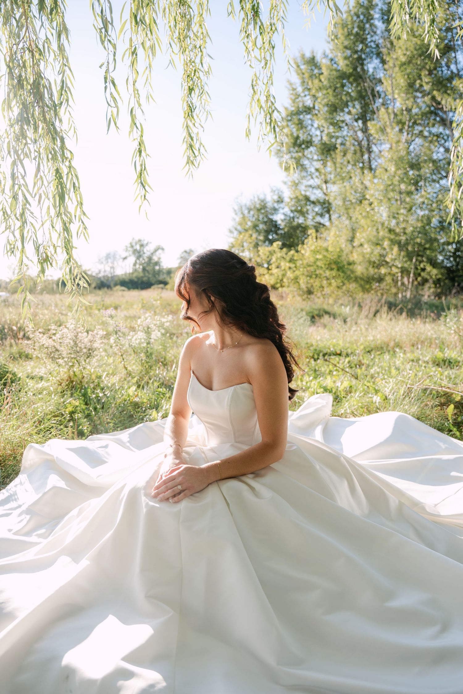 Bride in a strapless satin wedding gown sits in a sunlit meadow beneath willow branches, her dark hair pinned in a loose low updo.