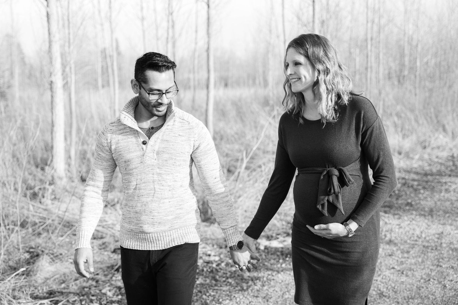 Expectant couple walking together on a snowy path while holding hands in a natural winter setting.