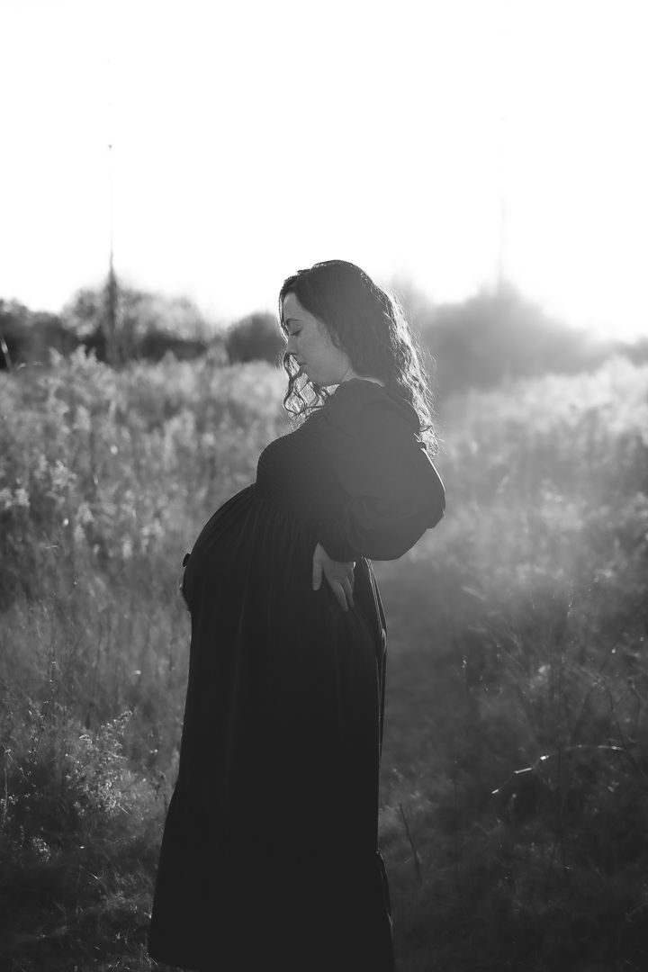 Silhouette of an expectant mother in a long dress standing in a sunlit field.