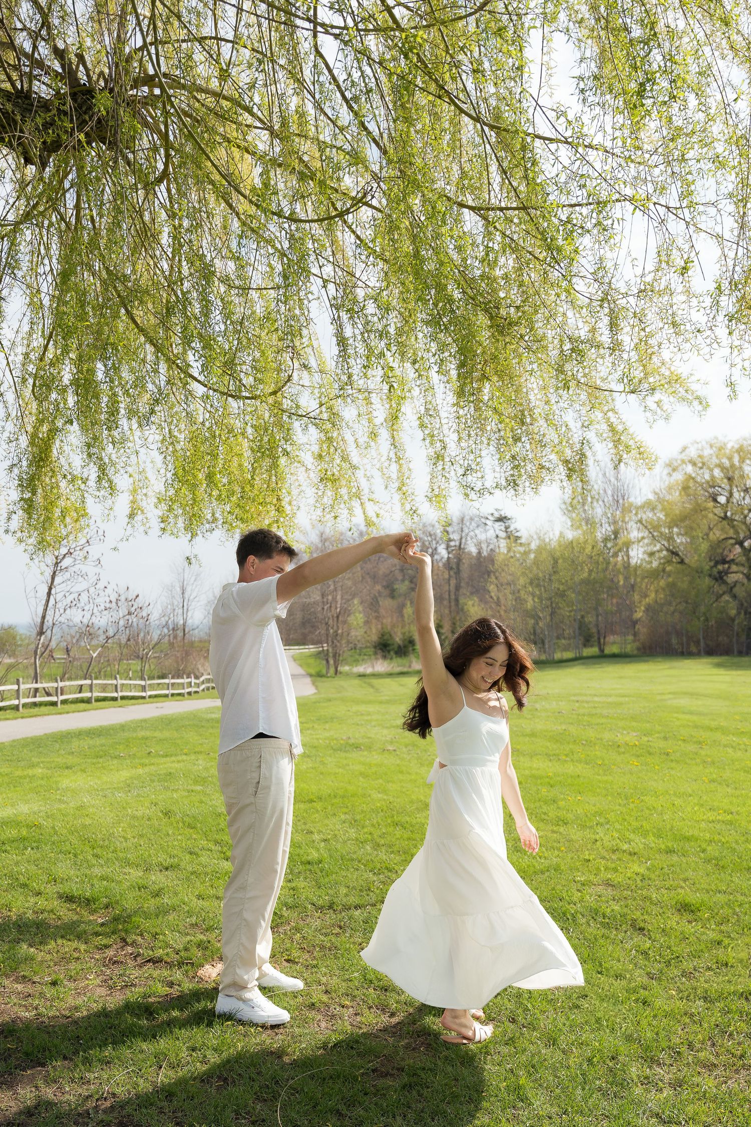 Couple dancing under a weeping willow tree on green grass, woman in white dress spinning, man in white outfit, sunny park.