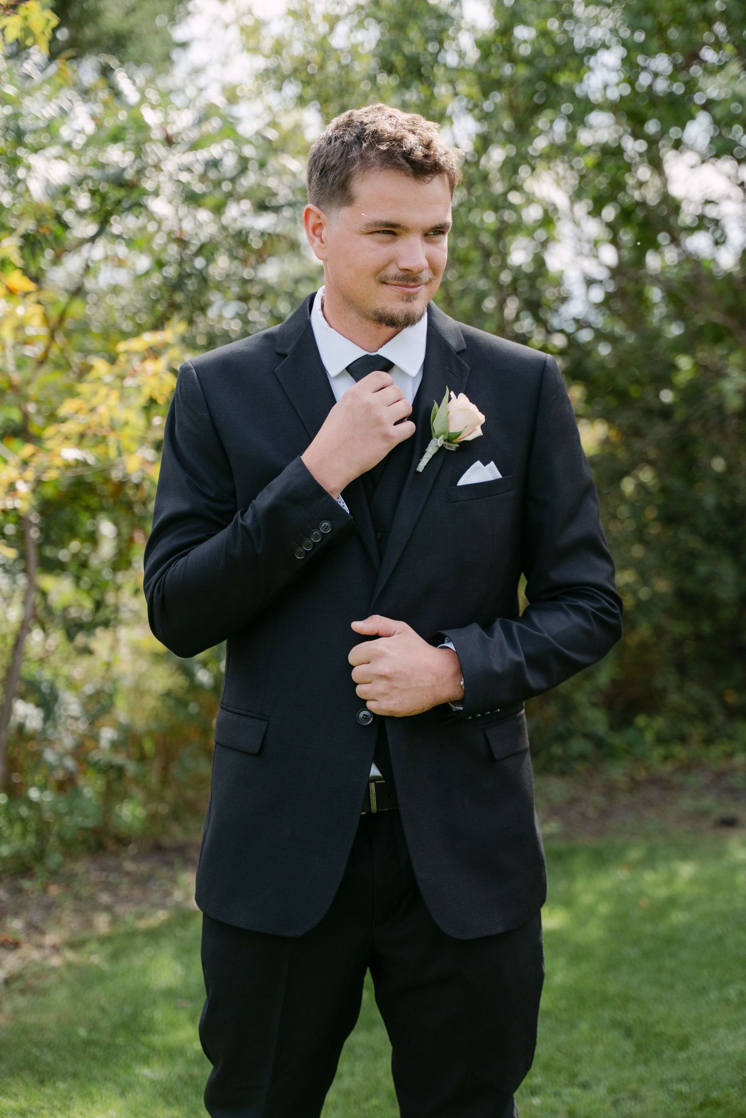 Groomsman in a black suit with a white rosebud boutonniere adjusts his tie while standing outdoors among green trees.