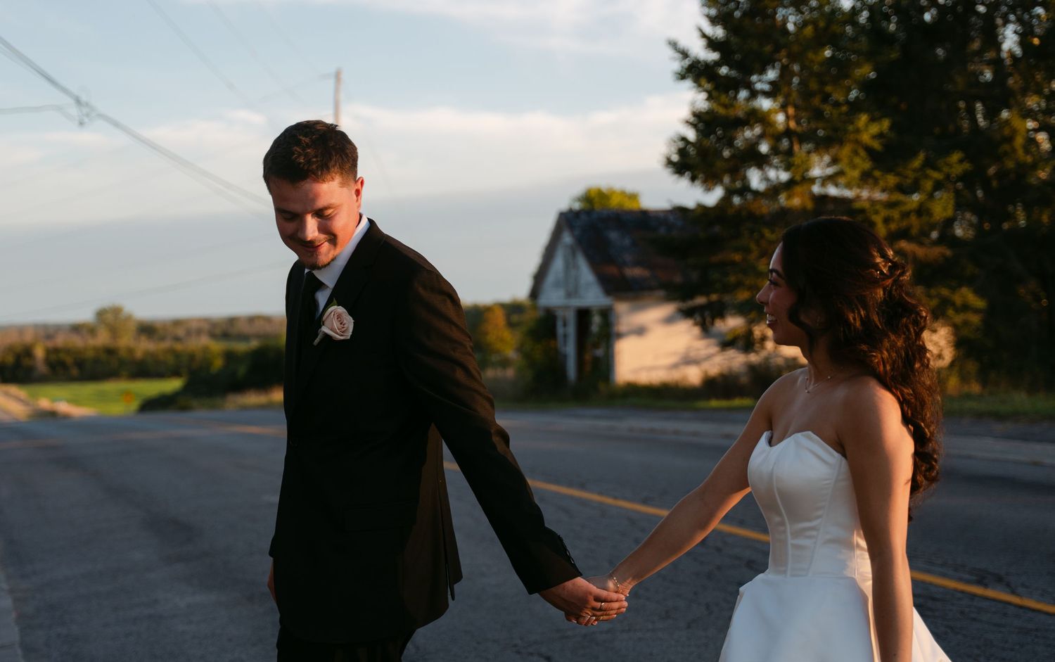 Smiling groom in a dark suit holds hands with a bride in a white wedding gown while walking down a country road at sunset.