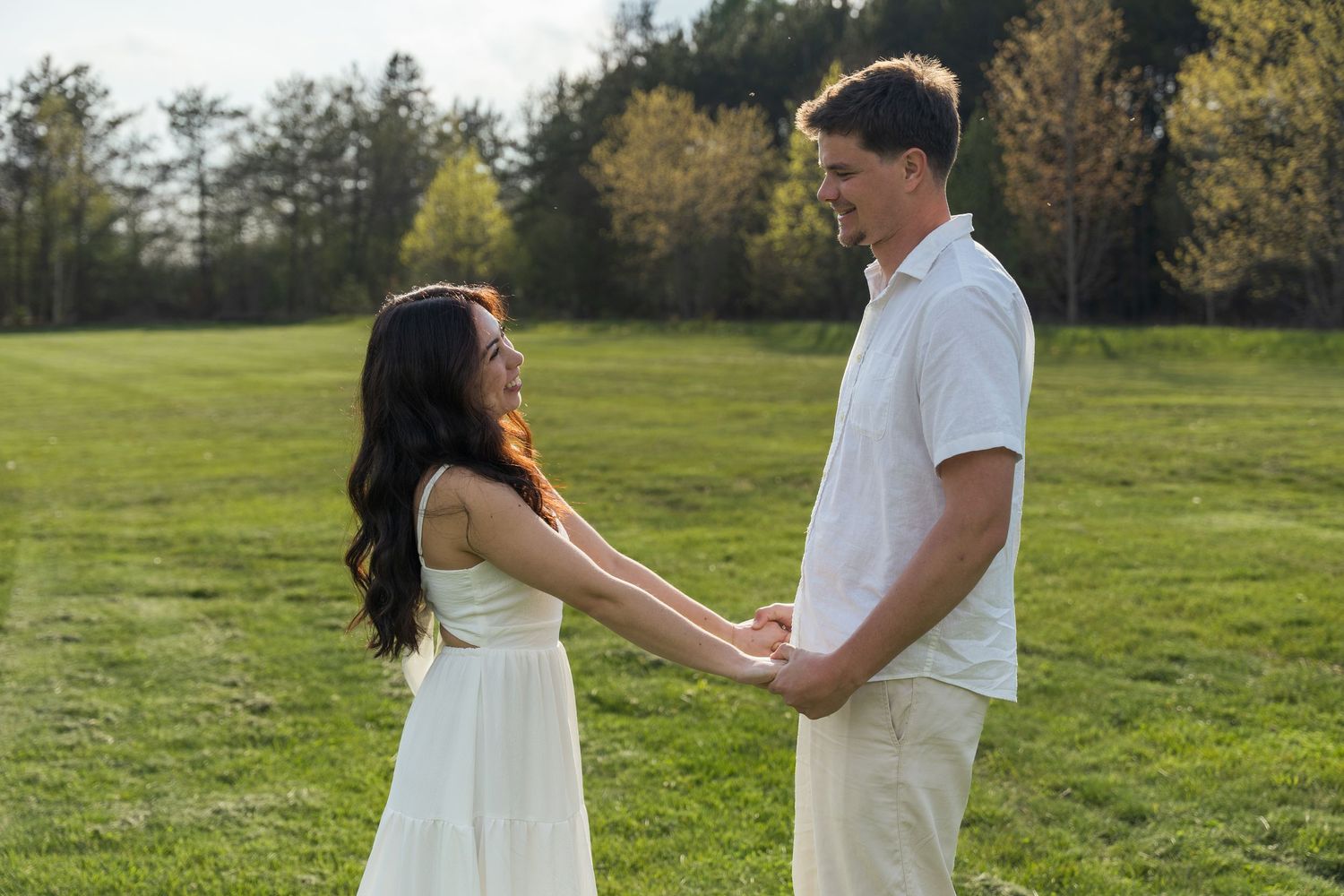 Couple in white outfits holding hands and facing each other in a sunny green grassy field with trees in the background.