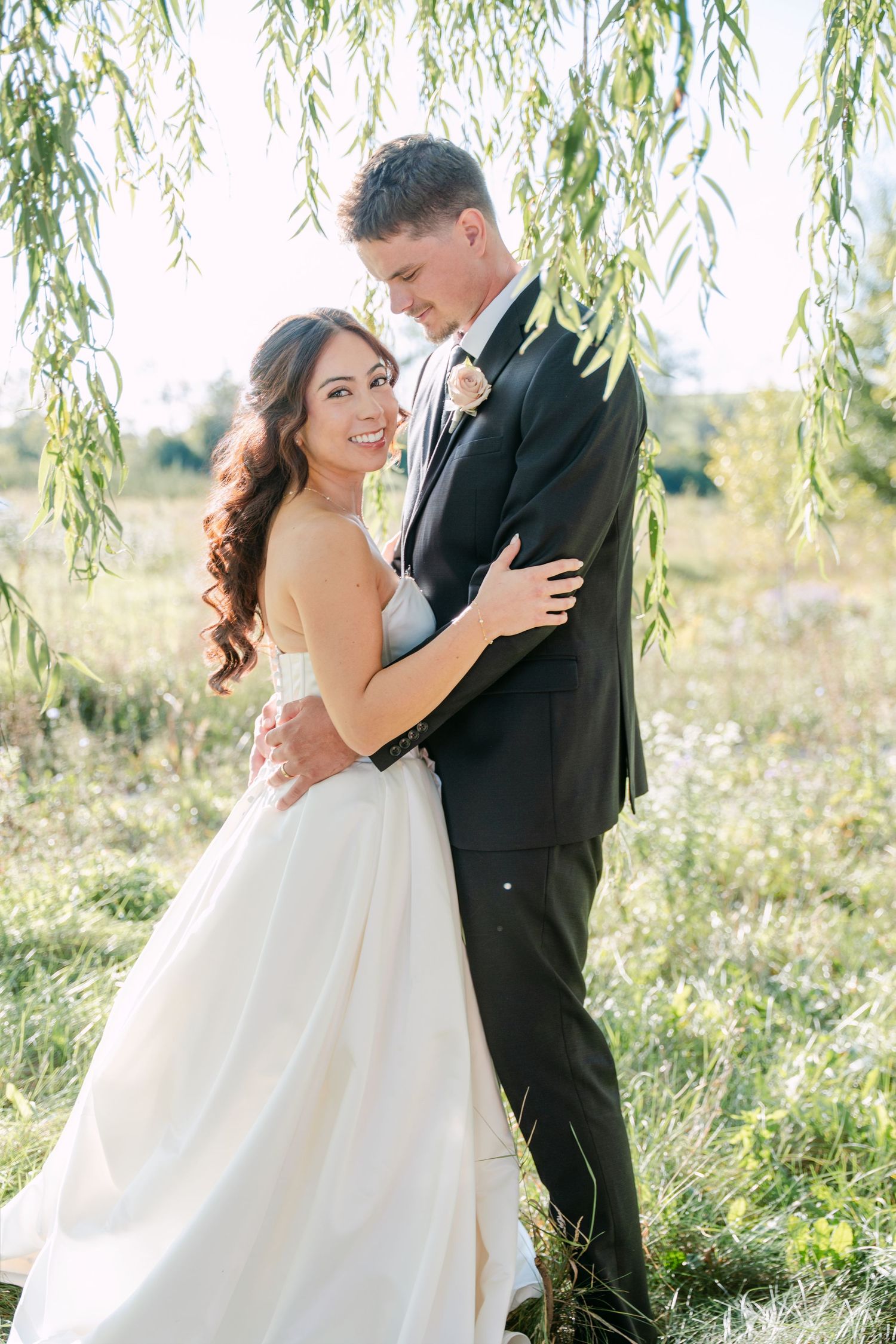 Bride in a strapless white gown embraces her groom in a black suit beneath willow branches in a sunny grassy field.