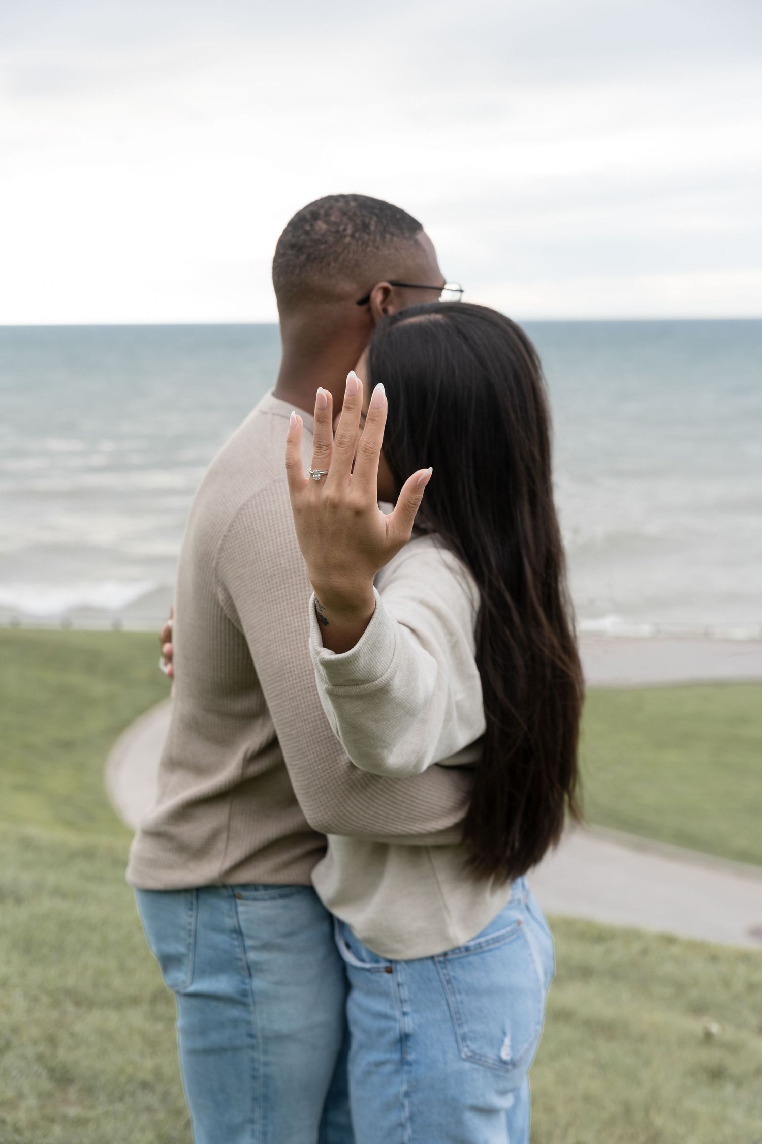Couple in matching sweaters embraces by the ocean while showing off an engagement ring.