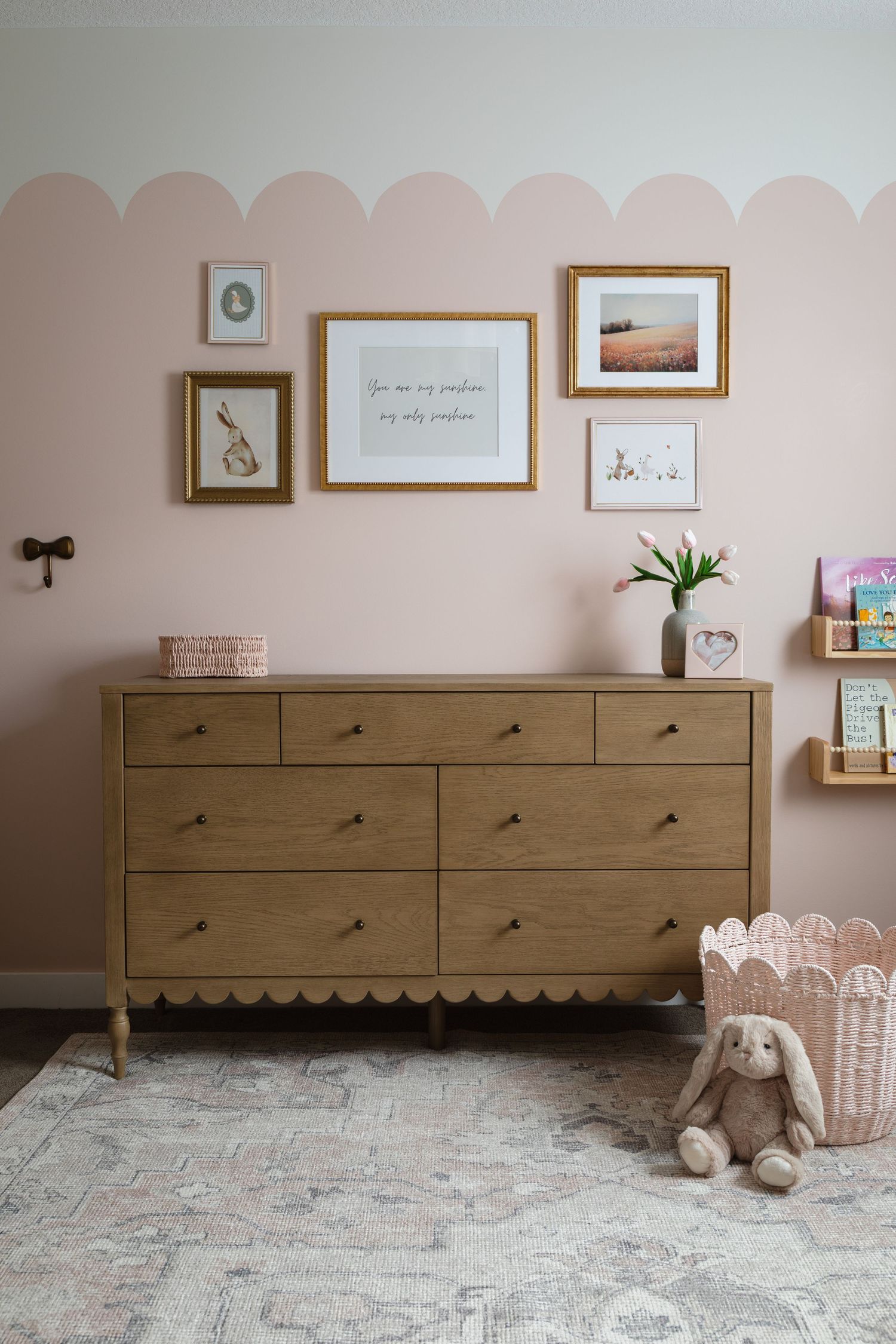 Pink nursery with scallop wall detail, wood dresser, gold-framed gallery wall, plush bunny toy, and floating book shelves.