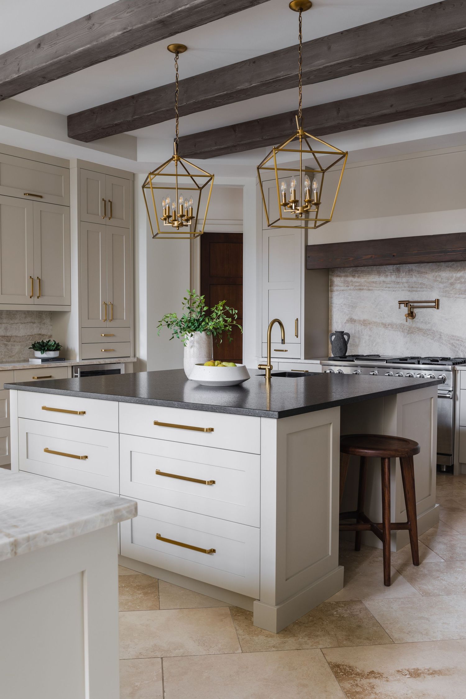 Classic kitchen island with dark countertop, brass pulls, gold lantern pendants, exposed wood beams, and travertine tile flooring.