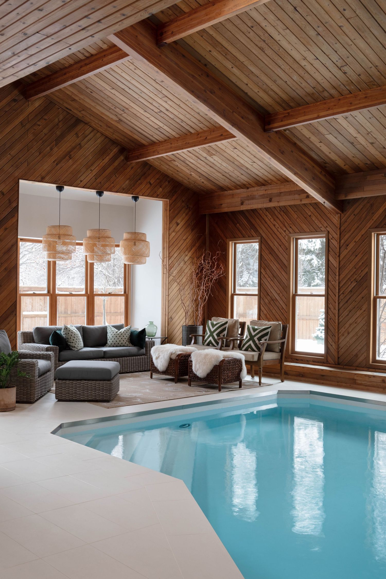 Luxury indoor pool room with vaulted wood ceiling, exposed beams, wicker pendant lights, and cozy lounge seating area.