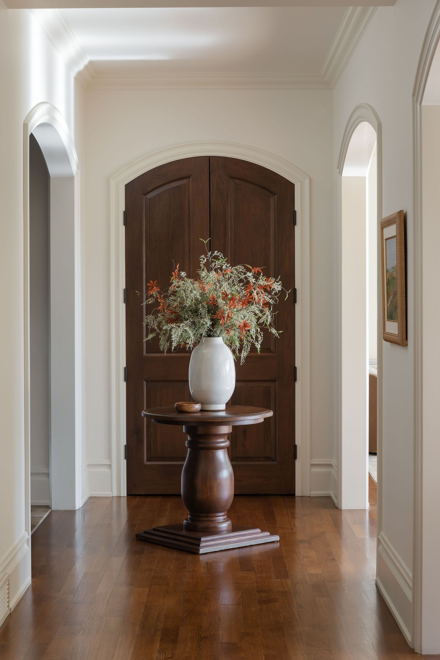 Classic foyer with dark arched wood doors, pedestal table, and white vase with floral arrangement.