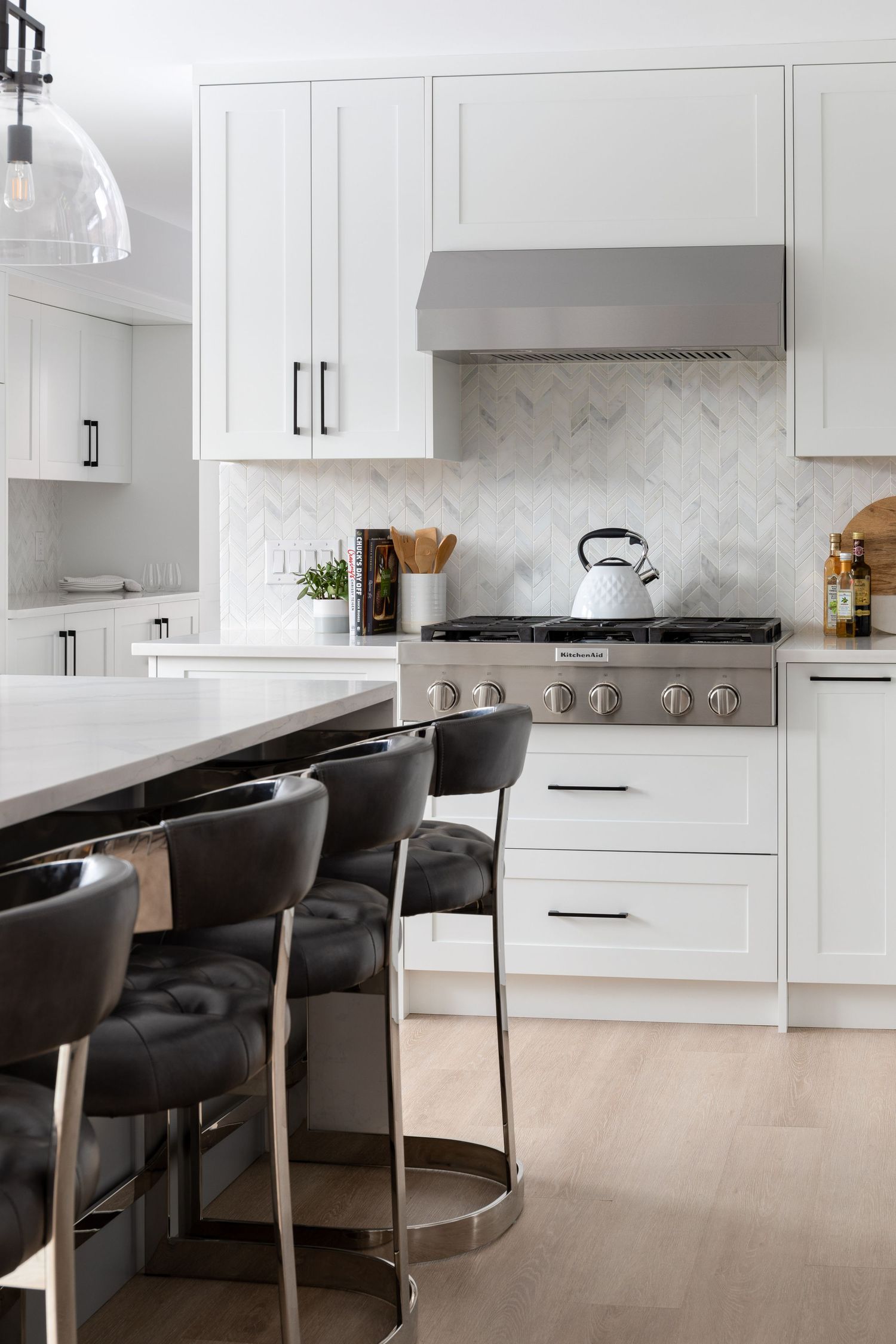 Elegant white shaker kitchen with marble herringbone backsplash, stainless range, black hardware, and leather bar stools at island.