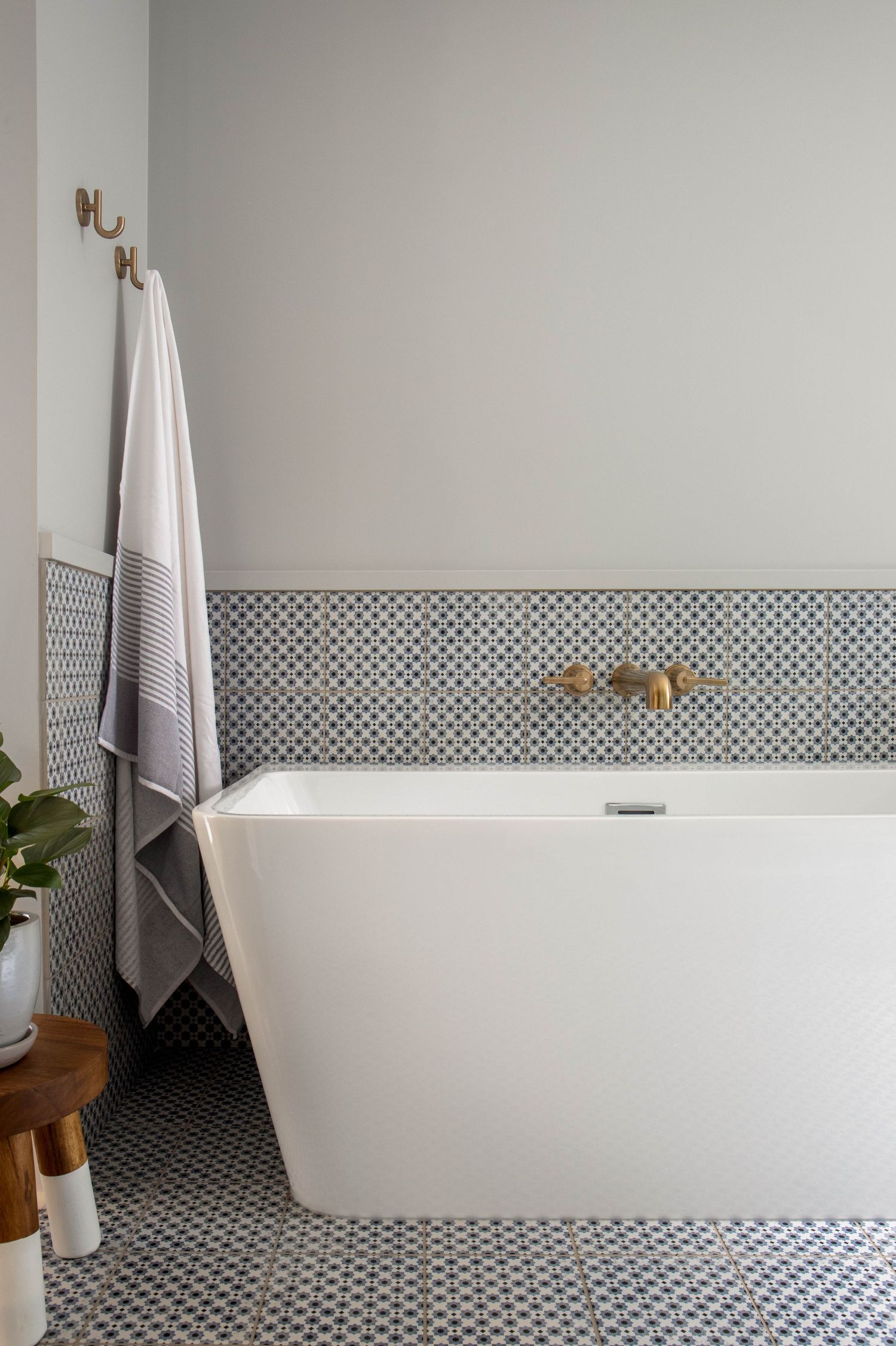 Freestanding white soaking tub with brass wall-mount faucet, patterned mosaic tile floor and wainscoting, and striped towels on hooks.