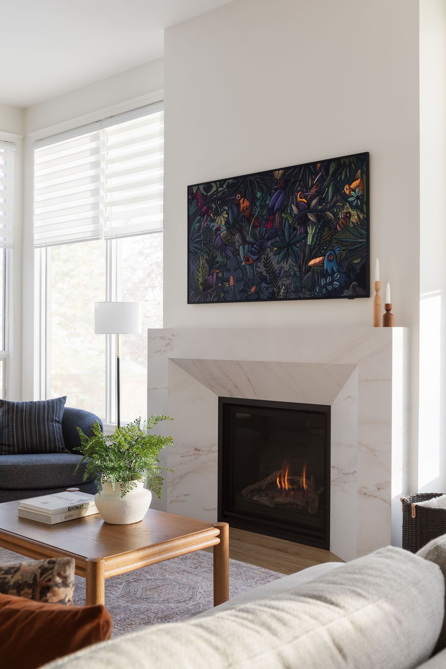 Modern living room with marble fireplace, colorful botanical art, navy sofa, and wood coffee table with green plant.