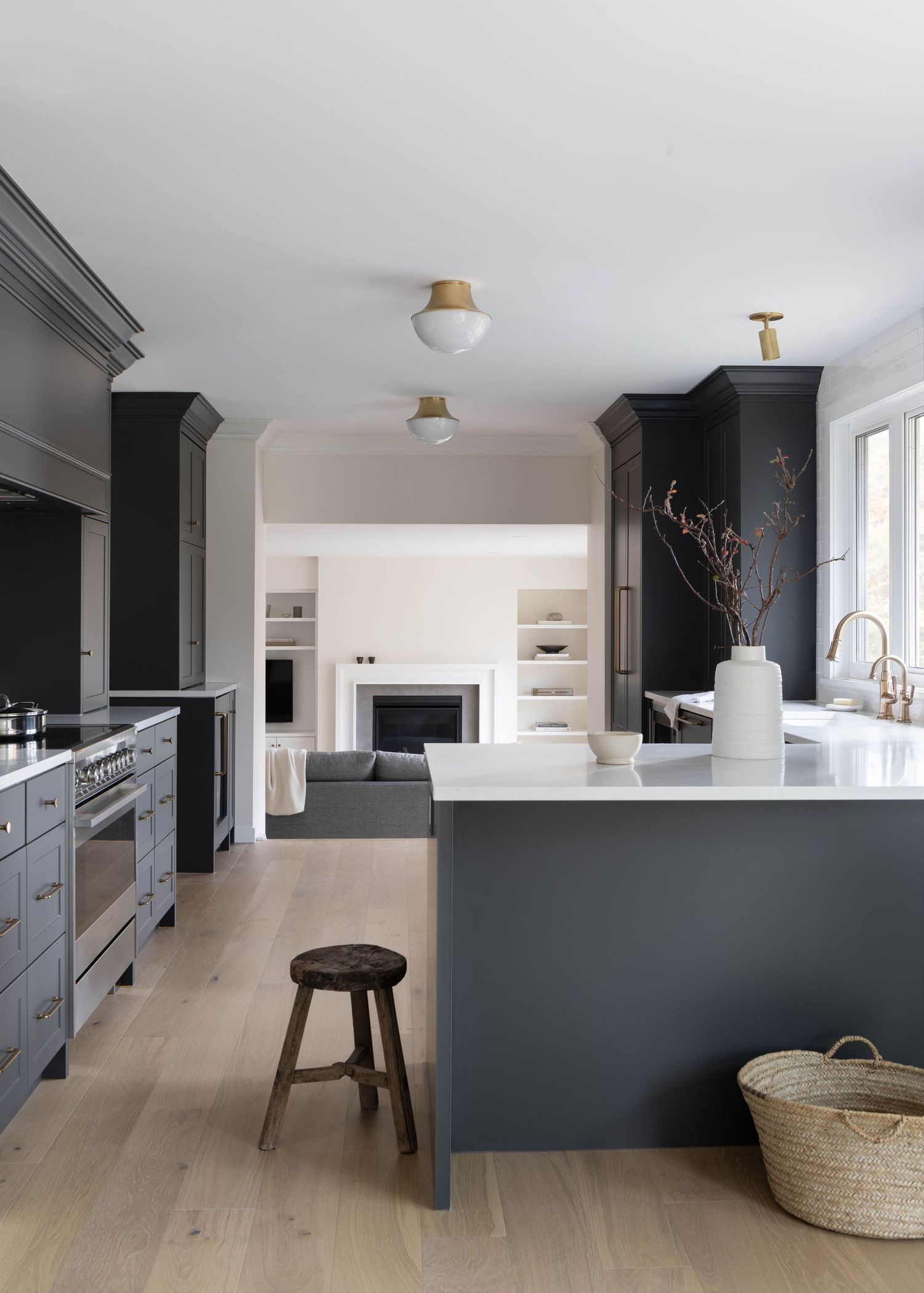 Contemporary dark gray kitchen with large island, light wood floors, brass accents, and view to living room fireplace.