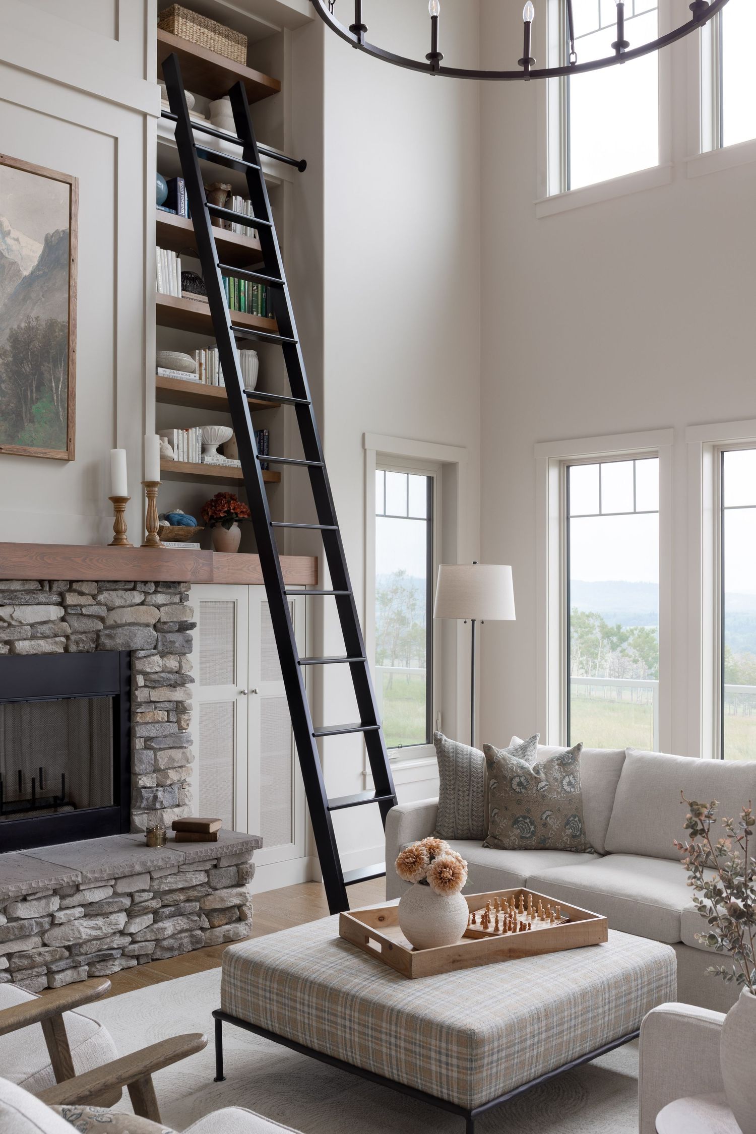 Grand living room with stone fireplace, tall library ladder, built-in shelves, plaid ottoman, and mountain views.