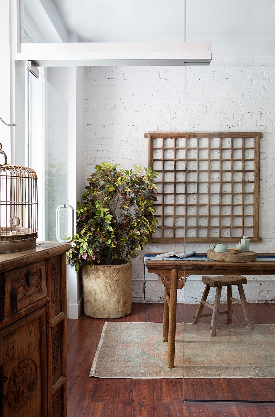 Eclectic interior with whitewashed brick, antique wooden furniture, lattice panel, birdcage, and tropical plant.