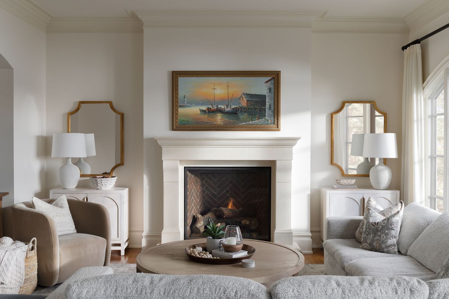 Elegant living room with white fireplace, gold mirrors, neutral sofa, and landscape painting above mantel.