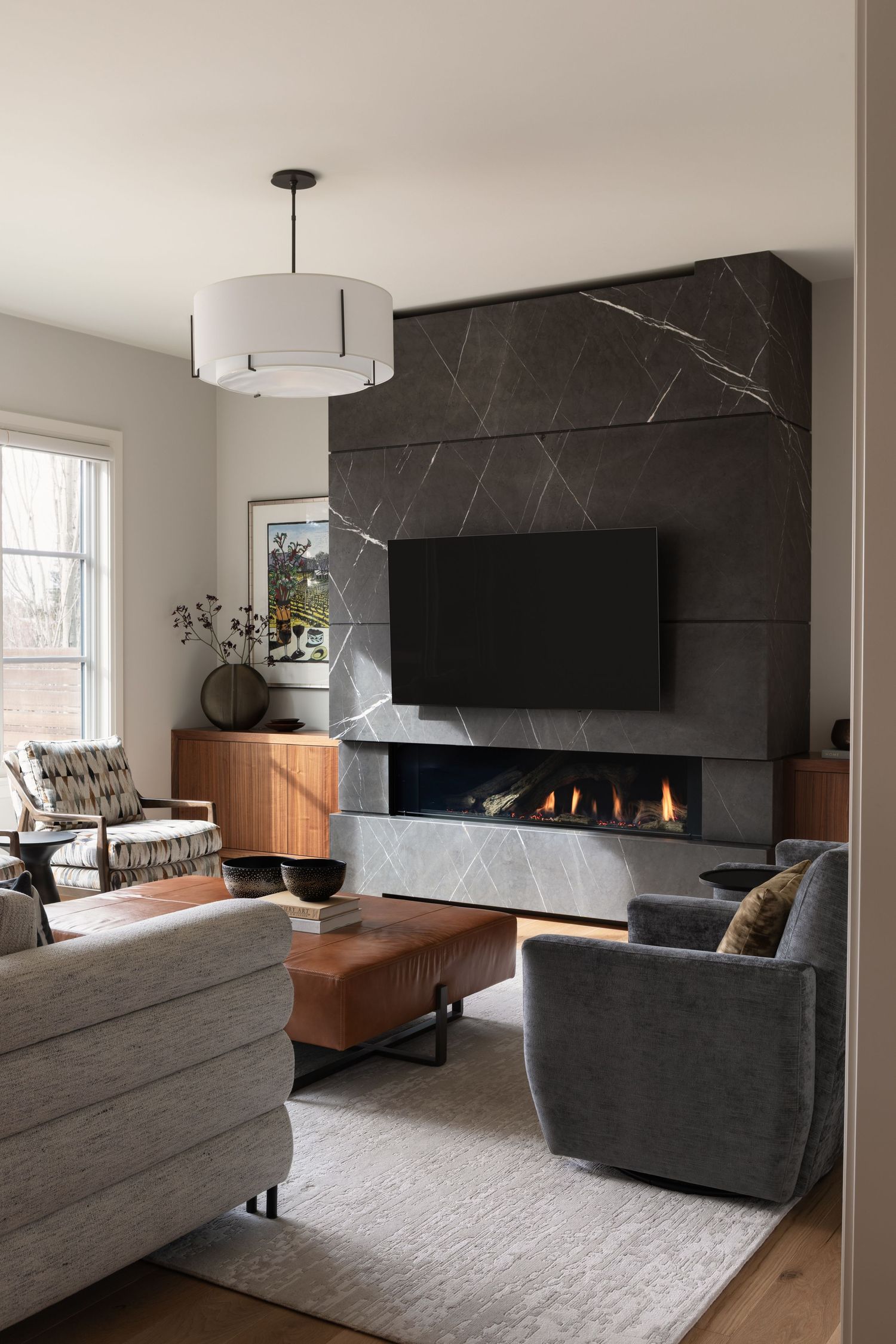 Modern living room with dark marble fireplace wall, mounted TV, gray velvet chairs, and leather ottoman on neutral rug.