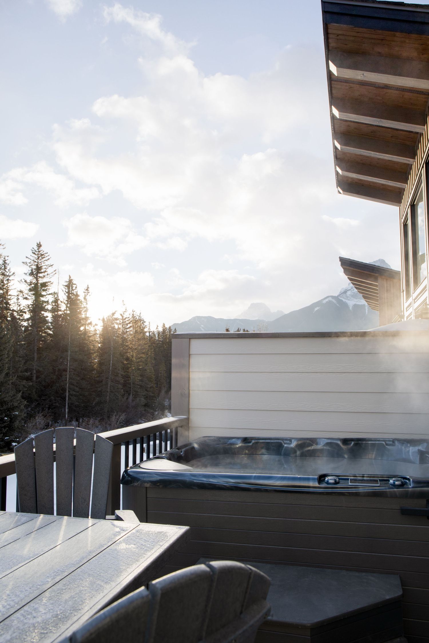 Steaming outdoor hot tub on snowy mountain deck with pine tree and mountain views at sunset.
