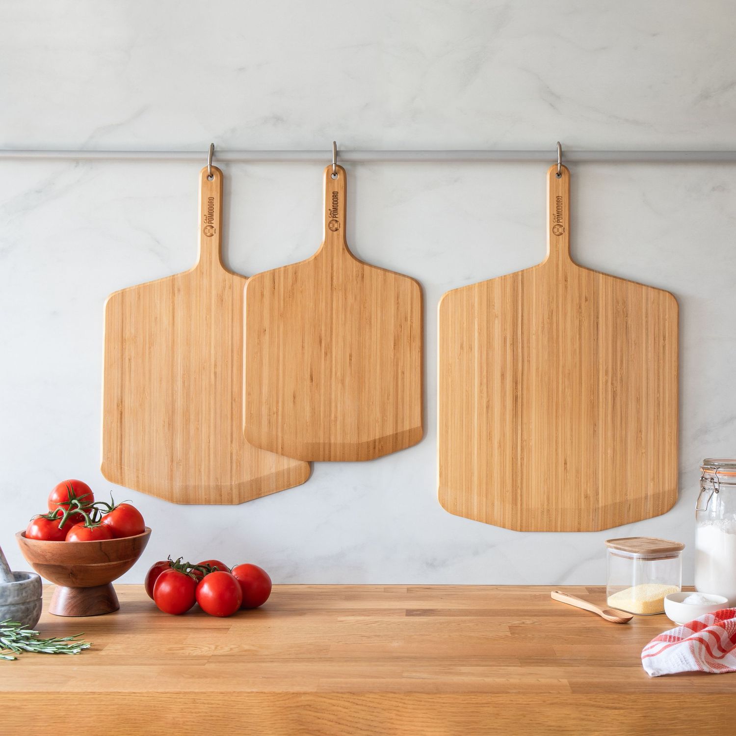Three bamboo pizza peel boards hanging on a marble wall above a wooden kitchen counter.