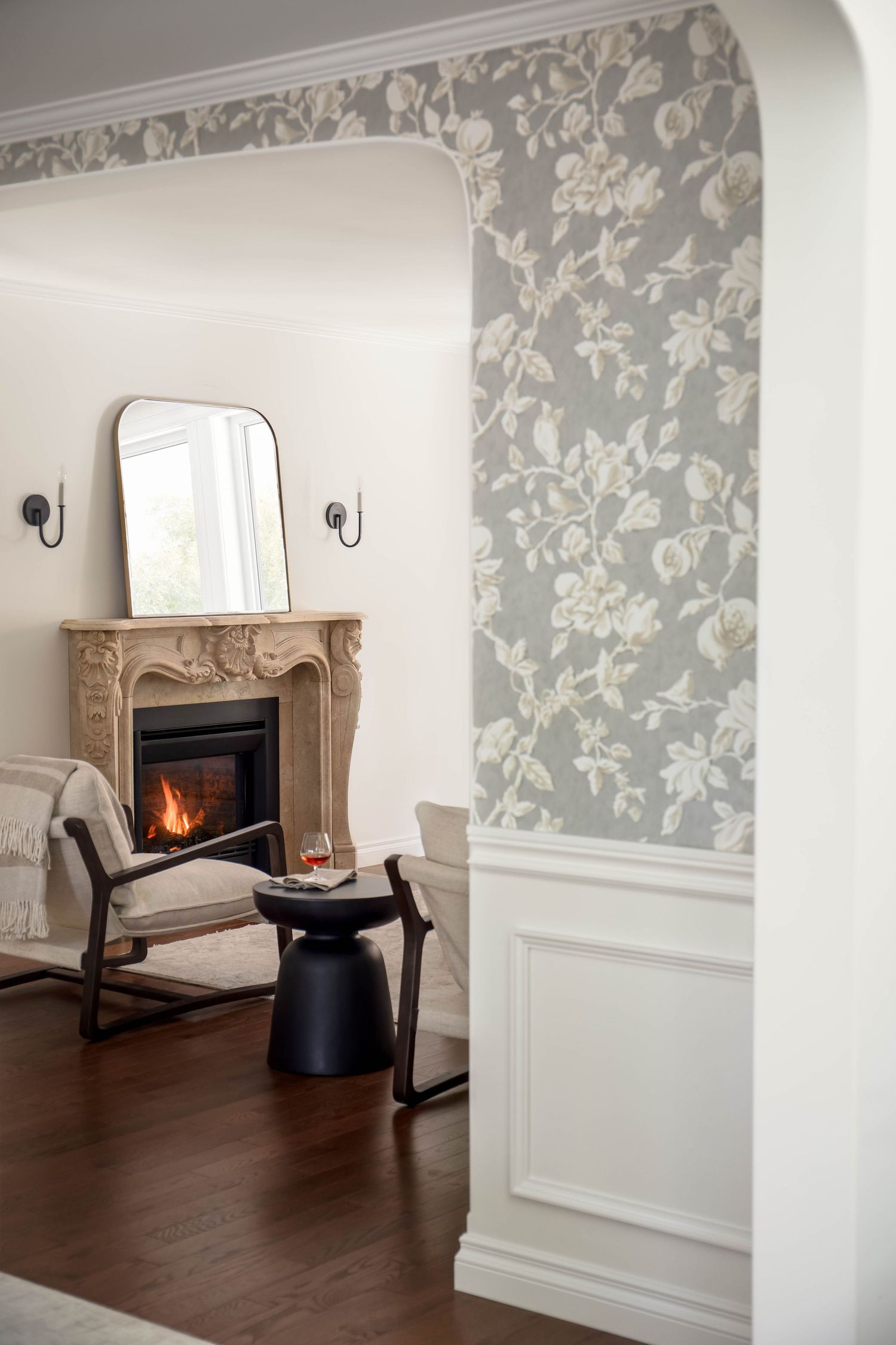 Cozy sitting room with ornate stone fireplace, floral wallpaper, rocking chair, and black side table.
