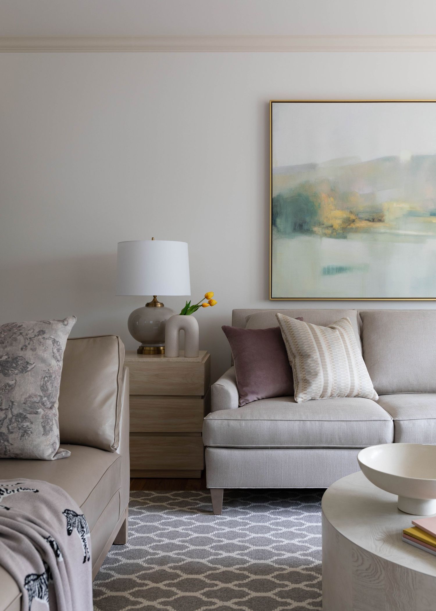 Serene living room with neutral sofa, landscape artwork, gray lamp, layered pillows, and geometric area rug.