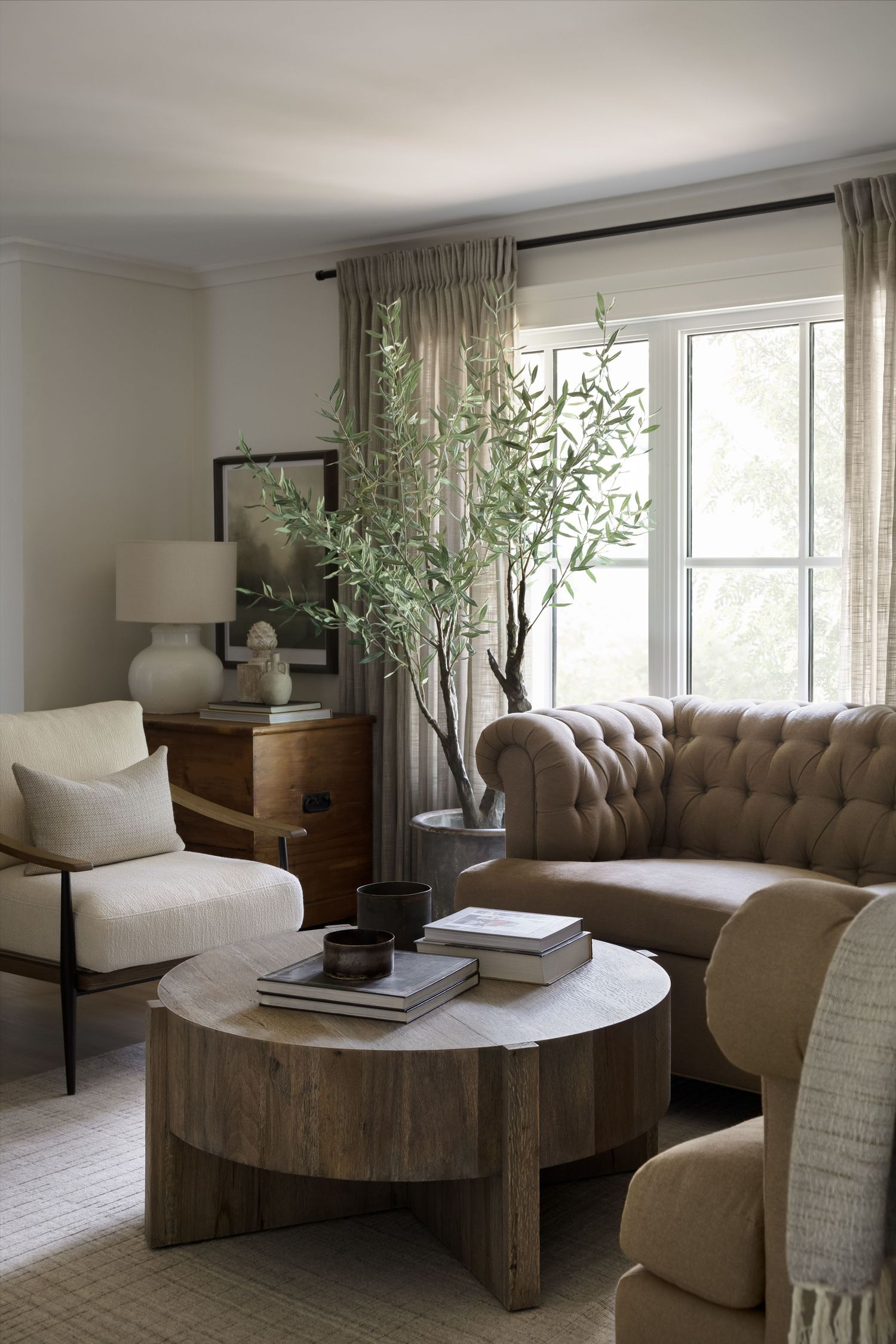 Cozy living room with tufted camel sofa, round wood coffee table, olive tree, and neutral linen armchair.