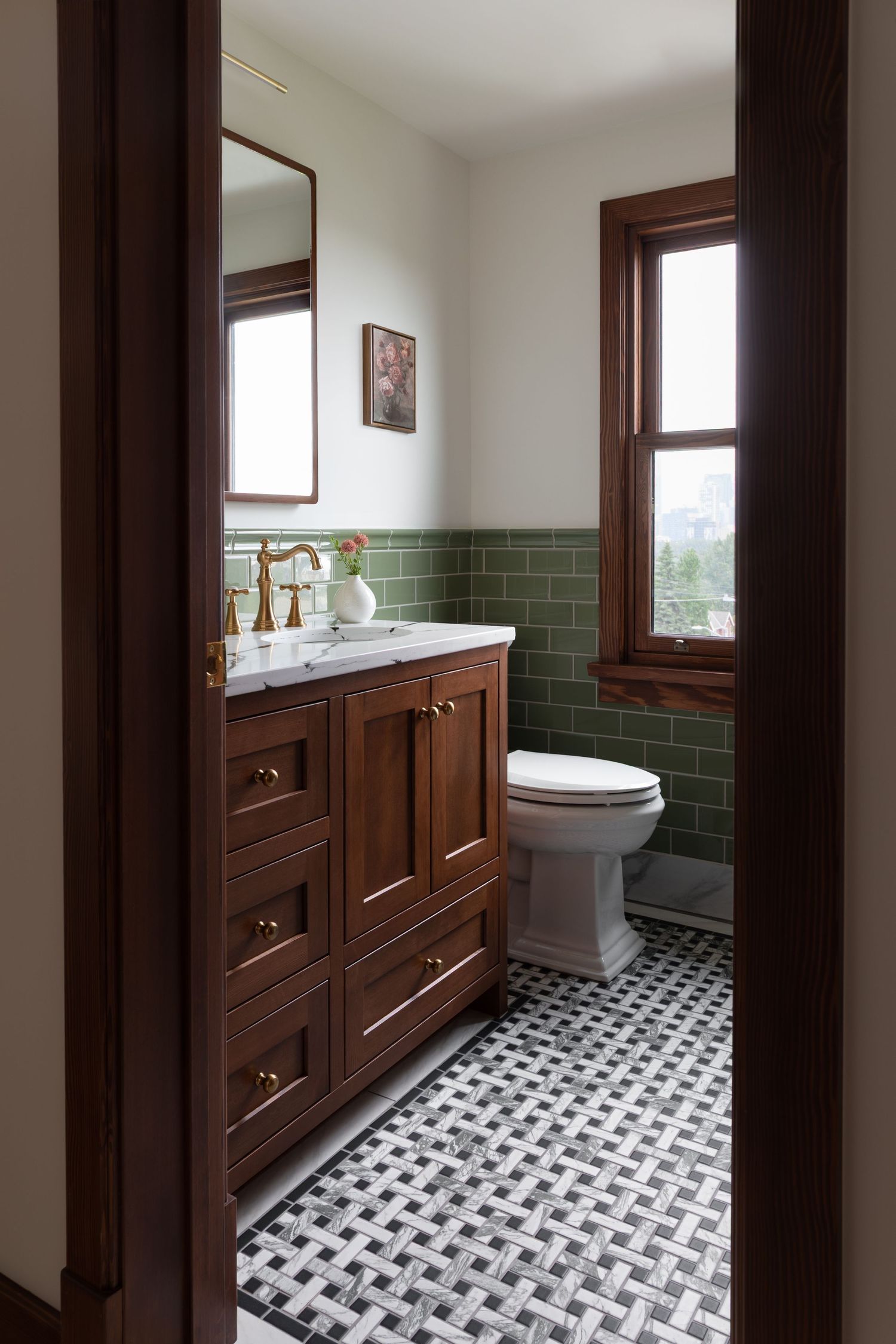 Elegant bathroom with walnut vanity, brass fixtures, green subway tile, geometric floor, and natural wood window trim.