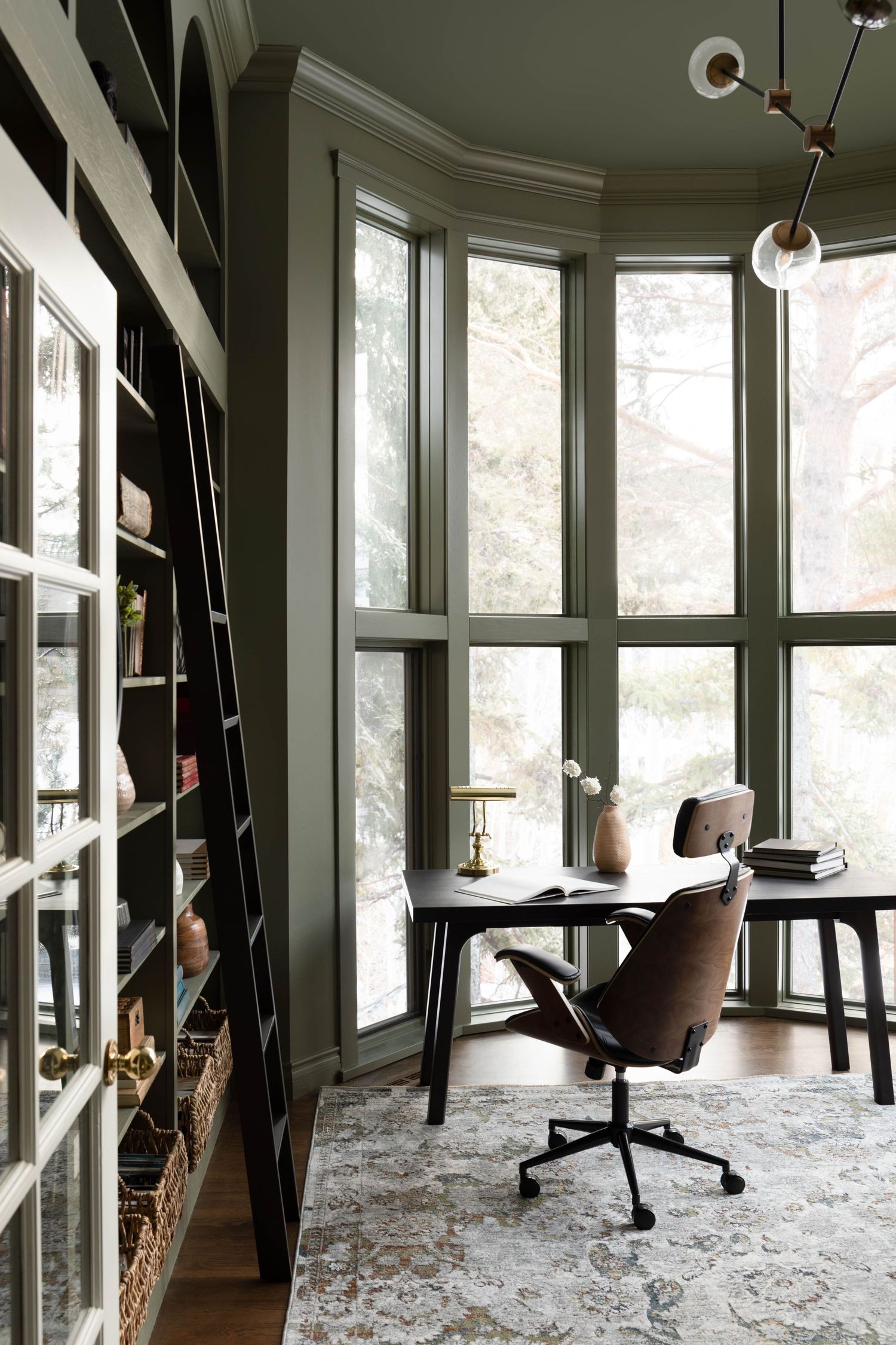 Sophisticated home office with sage green walls, floor-to-ceiling windows, built-in bookshelves, and leather desk chair.