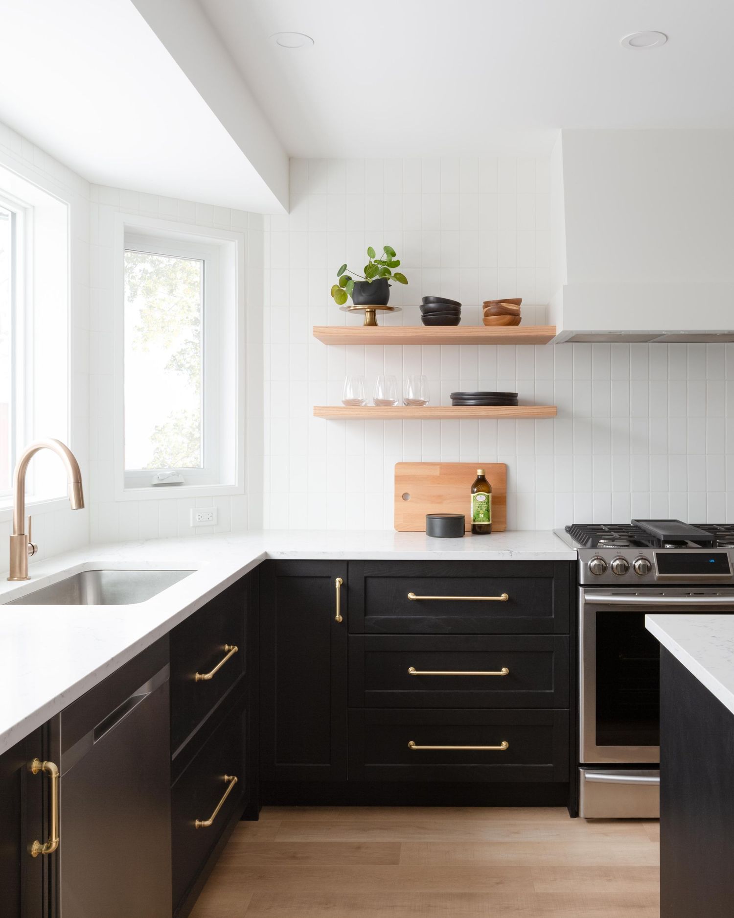 Modern kitchen with black lower cabinets, brass hardware, floating wood shelves, and white counters.