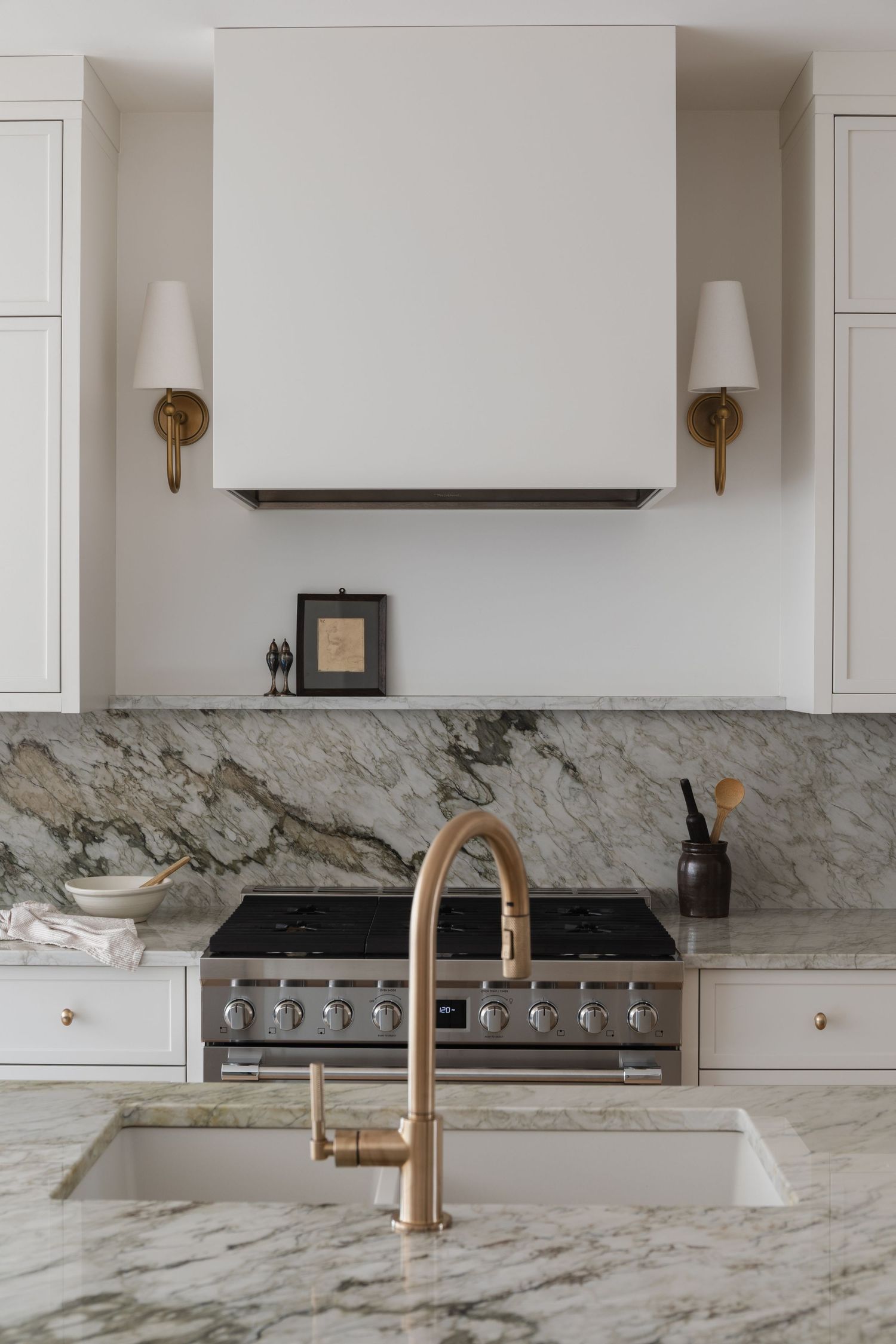 Elegant white kitchen with marble backsplash, brass faucet, range hood, and professional stainless steel stove.