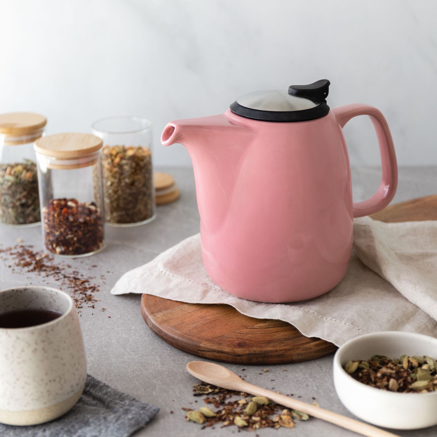 Pink ceramic teapot with black lid on a linen cloth, surrounded by loose leaf tea jars.