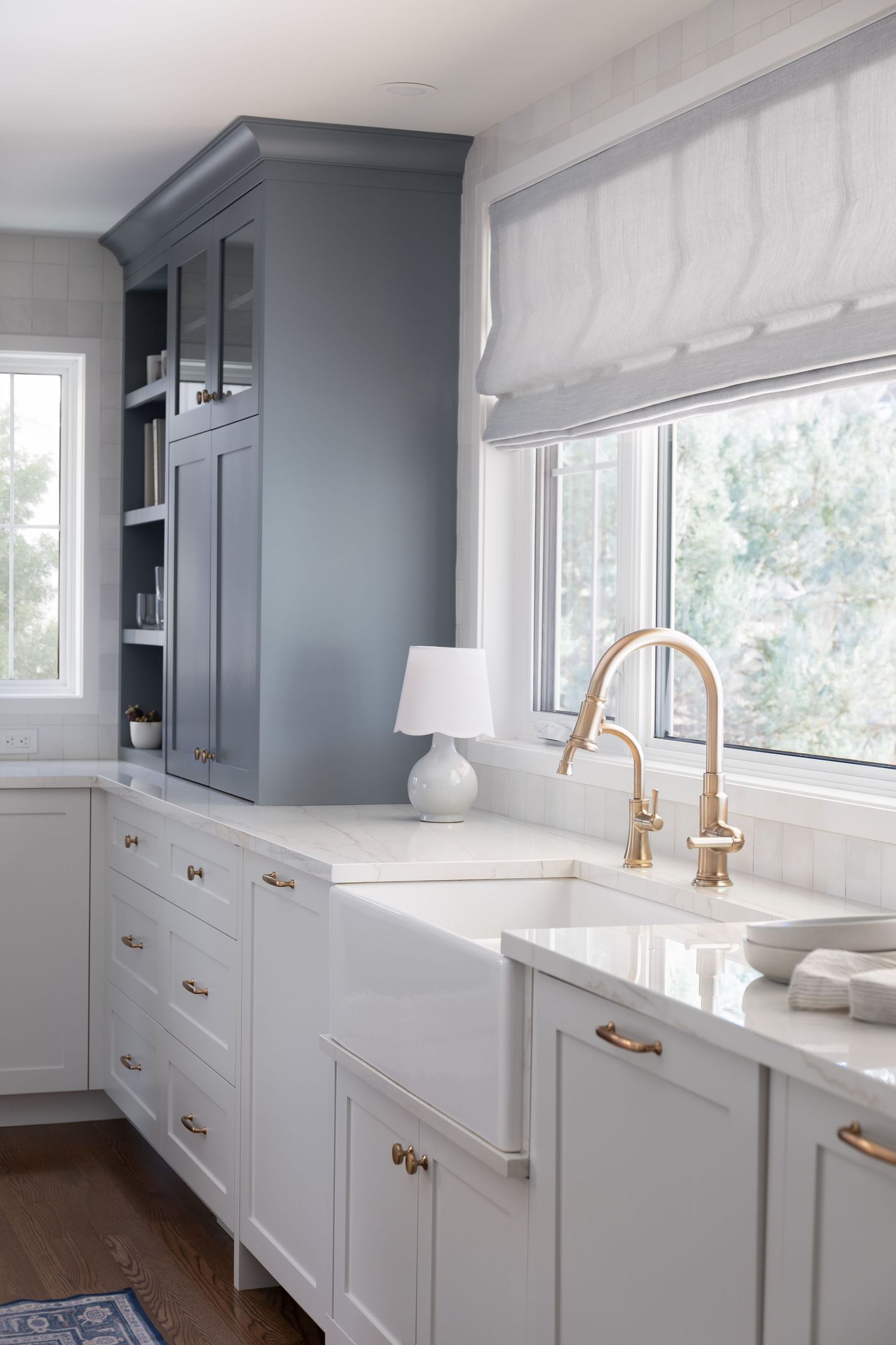 Classic kitchen with gray-blue cabinets, white farmhouse sink, gold faucet, brass hardware, and roman shade.