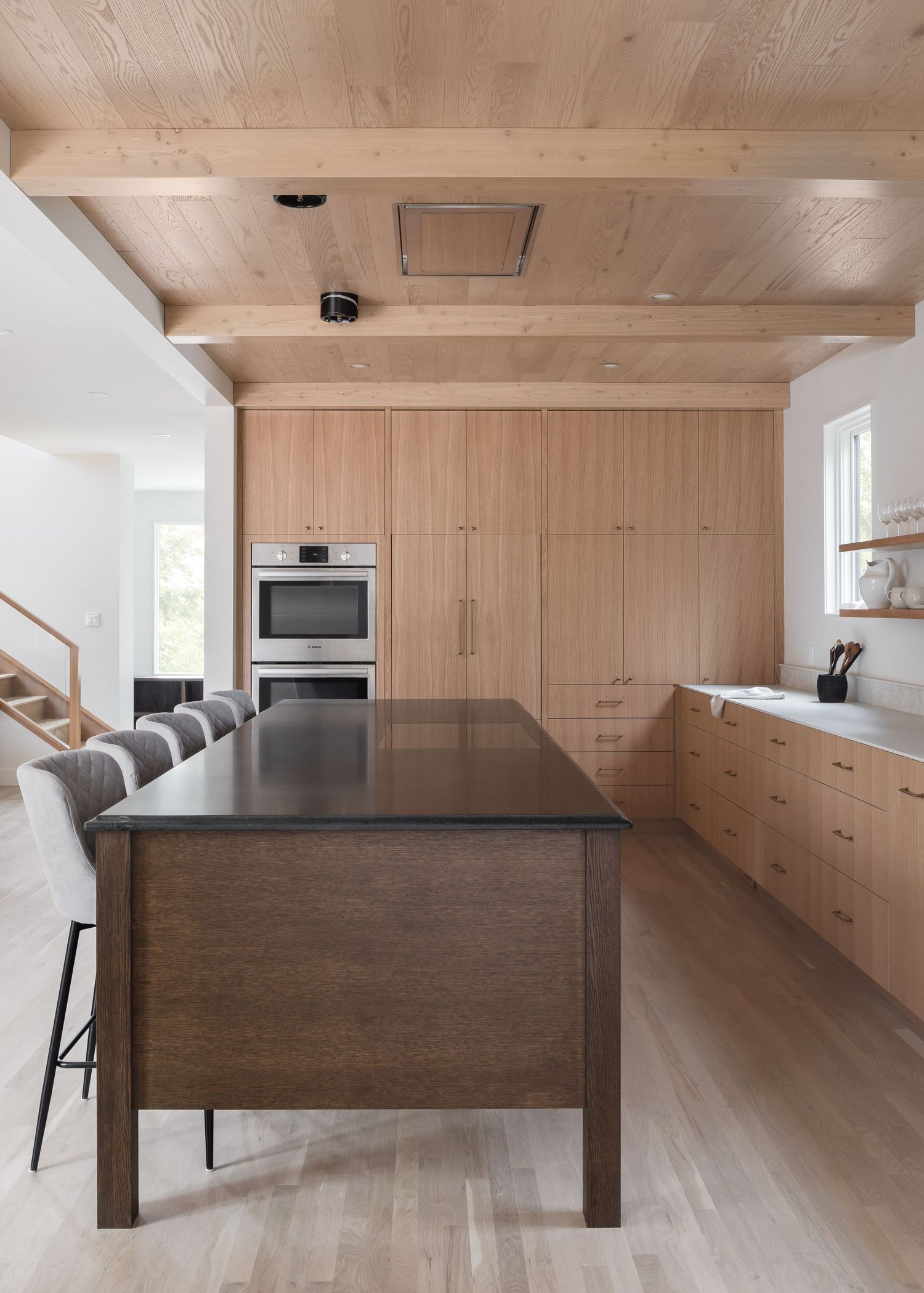 Minimalist oak kitchen with floor-to-ceiling cabinets, dark island, exposed wood beam ceiling, and stainless appliances.