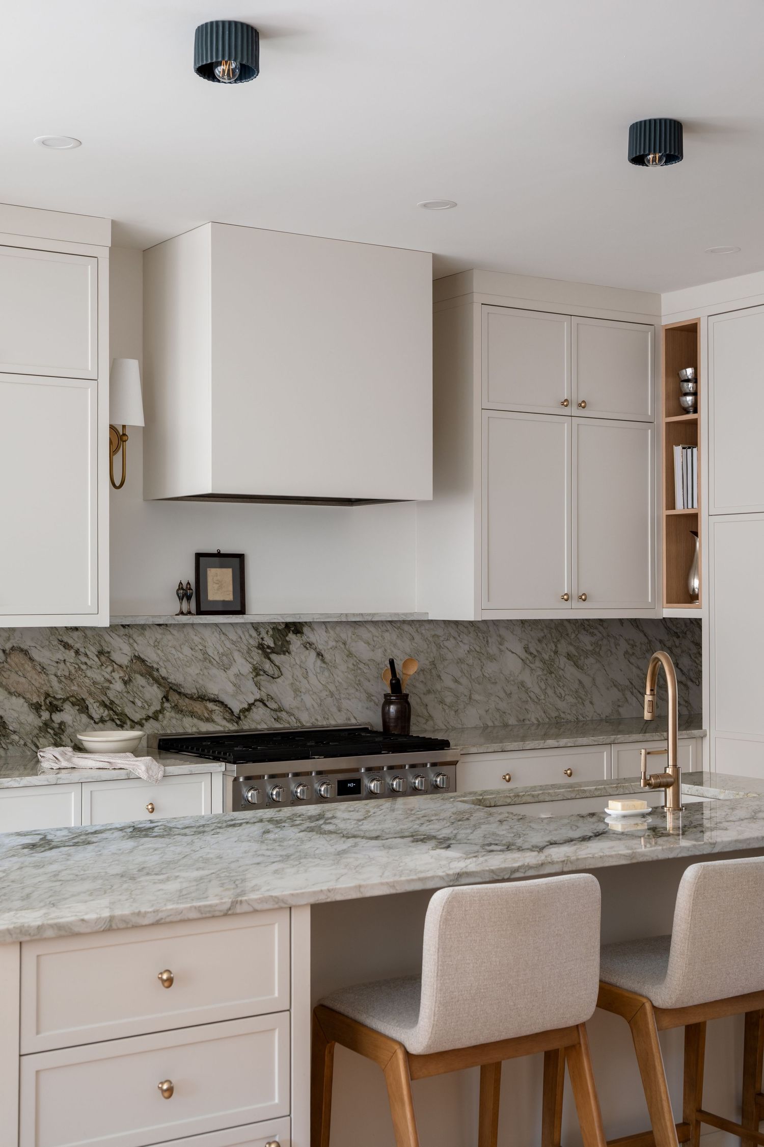 Luxury white kitchen with marble island, brass fixtures, upholstered bar stools, and custom range hood.