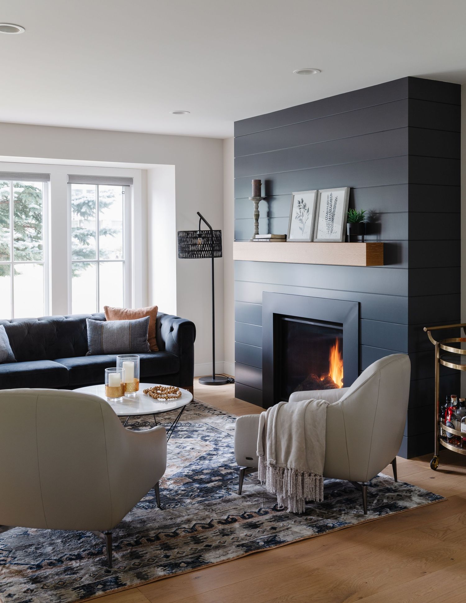 Modern living room with dark shiplap fireplace, cream accent chairs, navy sofa, and patterned area rug.