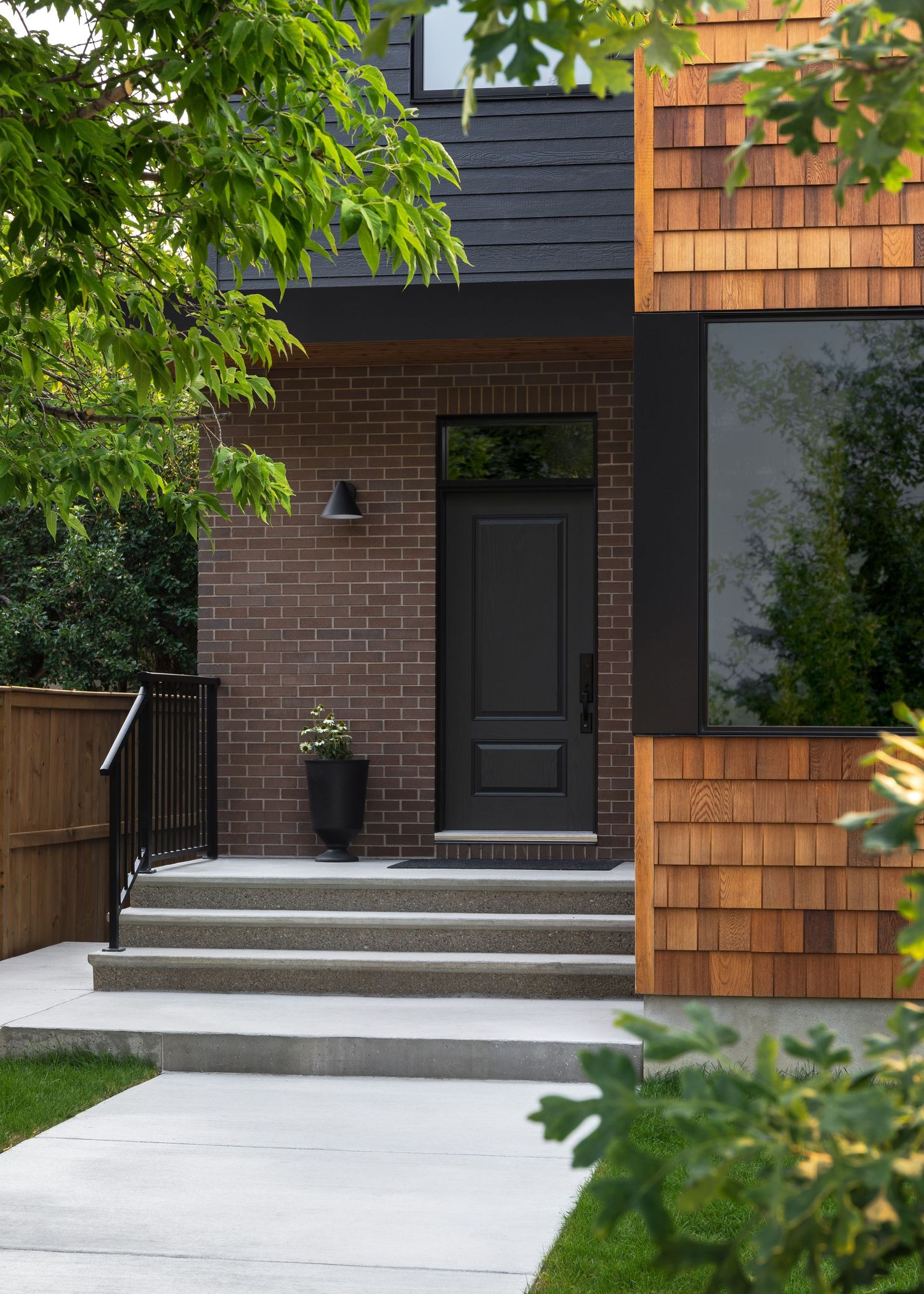 Modern home exterior with dark siding, cedar shingles, brick facade, black front door, and concrete steps.