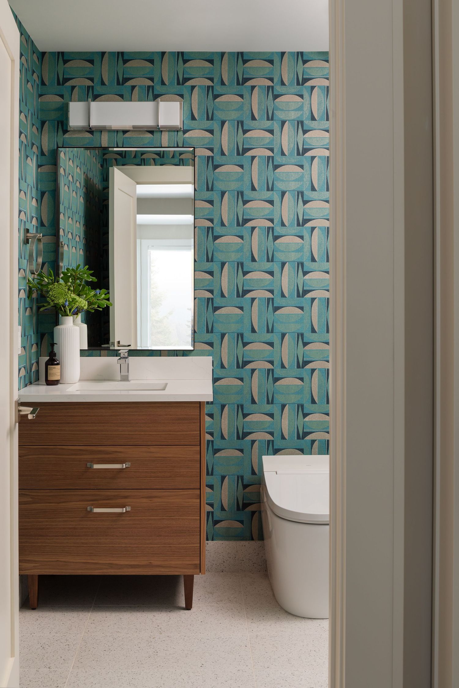Modern bathroom with teal geometric wallpaper, walnut vanity, white countertop, rectangular mirror, and sleek toilet.