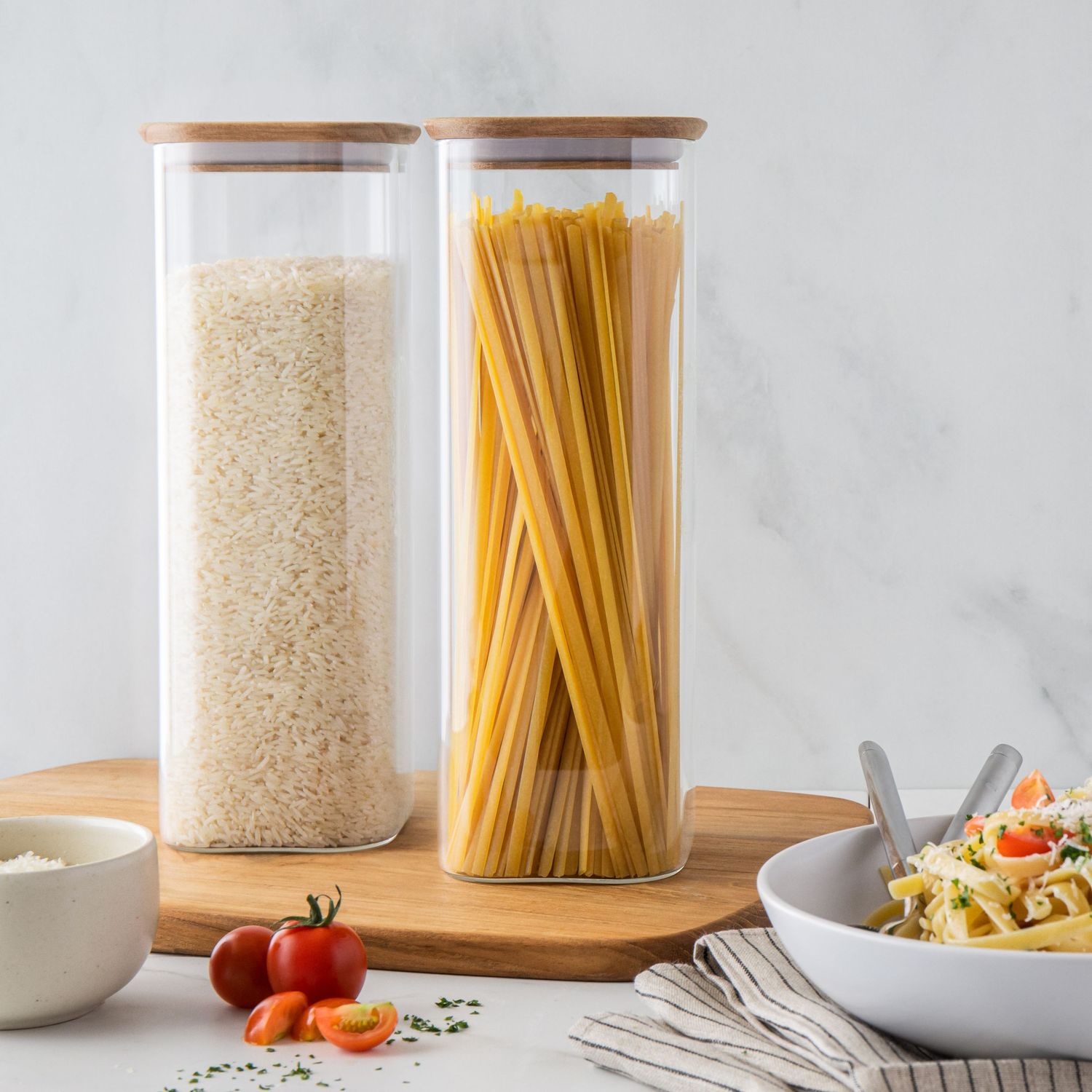 Tall glass storage jars with bamboo lids filled with rice and spaghetti on a wooden board.