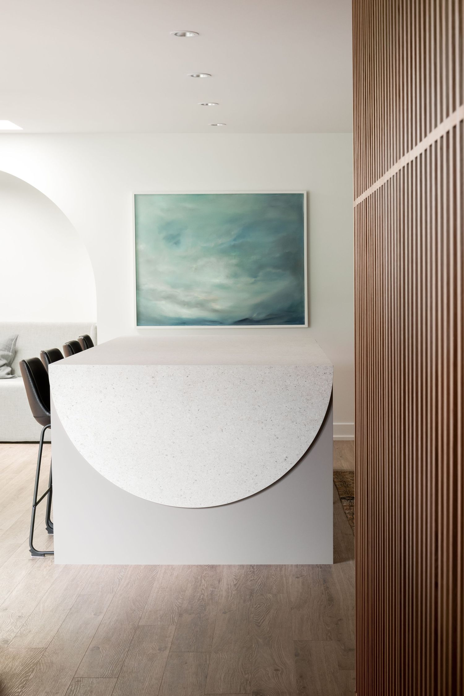Modern dining area with semicircular white terrazzo island, black chairs, teal artwork, and warm wood slat wall divider.