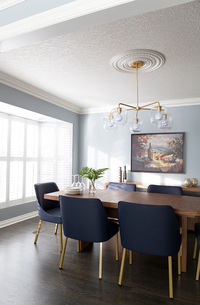 Contemporary dining room with navy velvet chairs, gold legs, wood table, brass chandelier, and blue-gray walls.