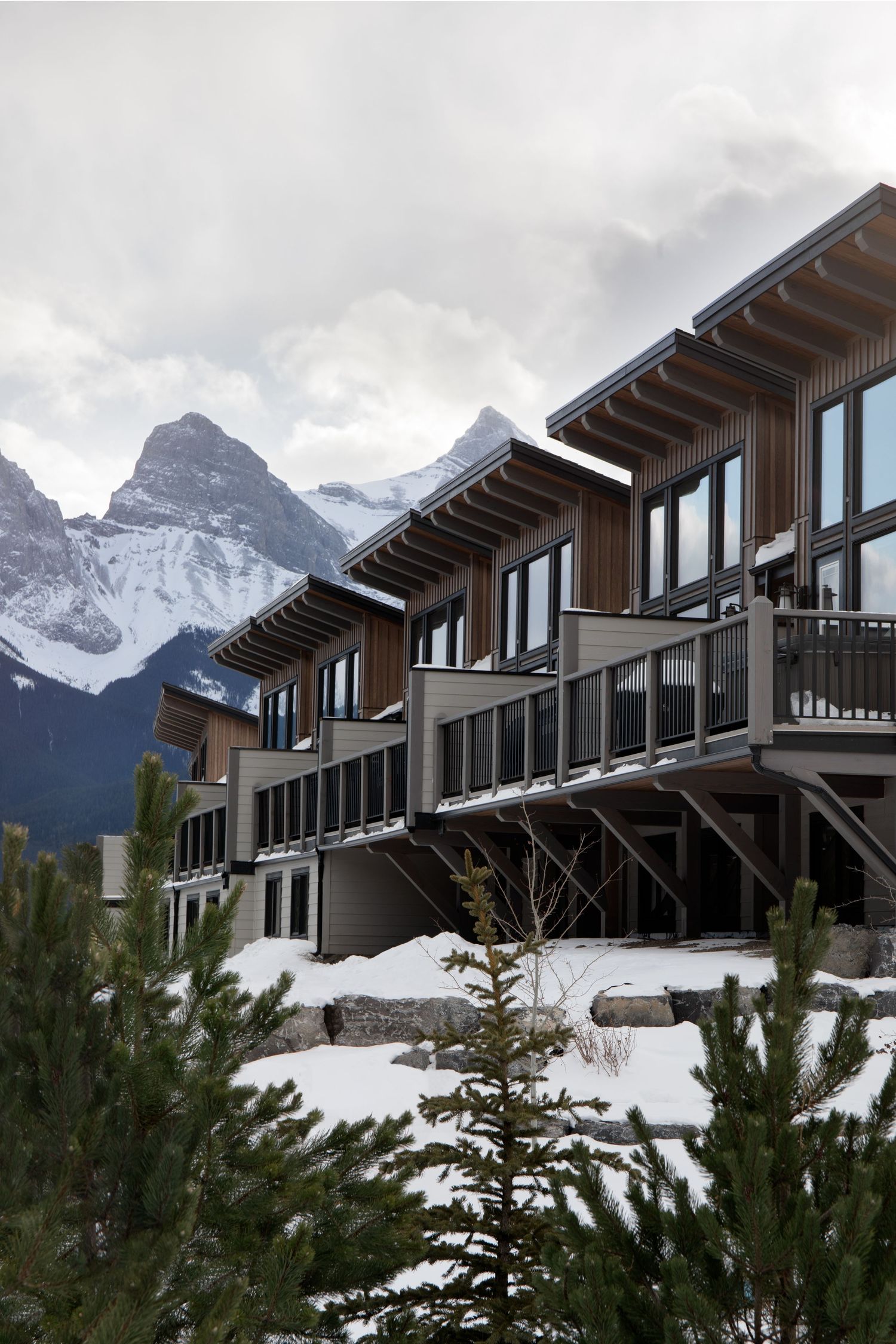 Modern mountain chalet with wood and glass facade, snow-covered landscape, and dramatic Rocky Mountain peaks backdrop.